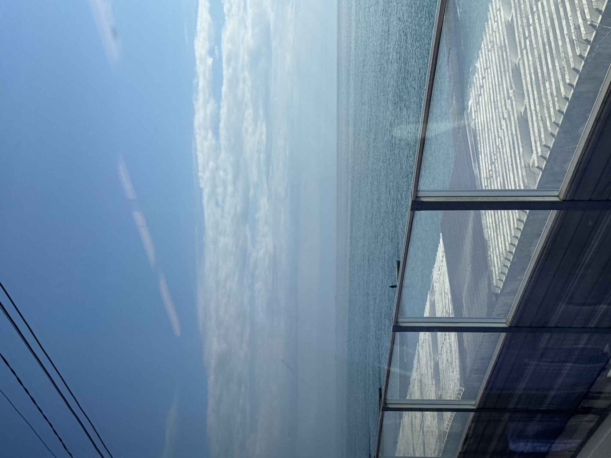 The image depicts a view from a high vantage point, likely inside a building, overlooking the Black Sea in Sochi, Russia. The frame is dominated by the reflection of what appears to be the interior ceiling or roof structure, visible through the glass windows. In the distance is the sea, with a visible horizon and a sky covered by patchy, light clouds. Some power cables are visible in the image. The image is taken at an oblique angle, creating a slightly distorted perspective. The photograph's composition is split between the interior architecture and the natural seascape.