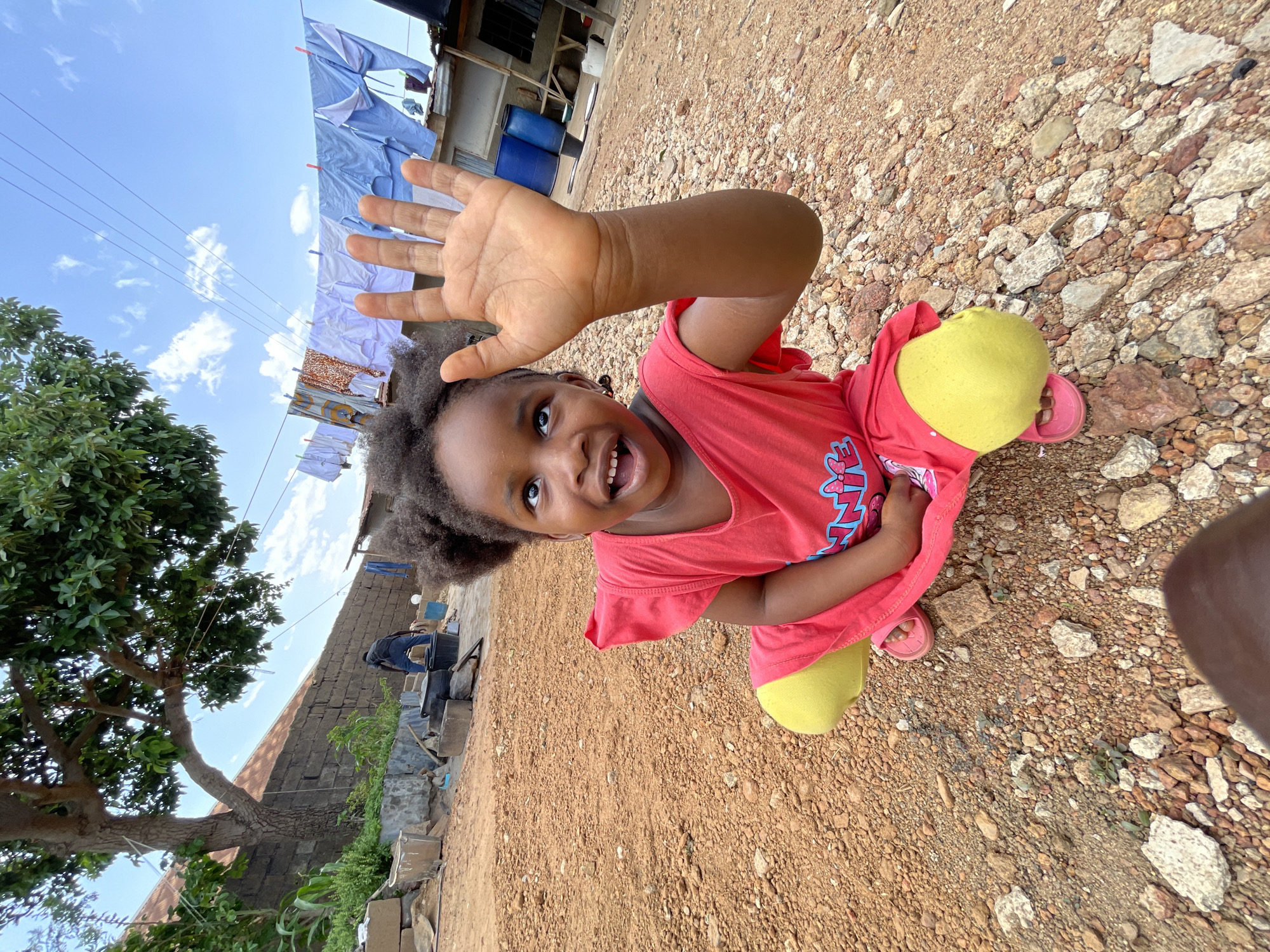 A young Black girl in a red shirt and yellow leggings is kneeling on a reddish-brown dirt and rock surface, holding her hand up towards the camera. She is smiling, and her gaze is directed slightly upwards. A tree with green leaves is visible in the lower left of the image. Clothes are hanging on a line in the background. A wall is visible behind the clothesline, and what appears to be a blue barrel is partially visible above and to the right of the clothesline. Based on the environment and the style of building, the image appears to be taken in an urban or semi-urban location. The city cannot be definitively confirmed from the image alone, but the prompt suggests the location is Jos, Nigeria.