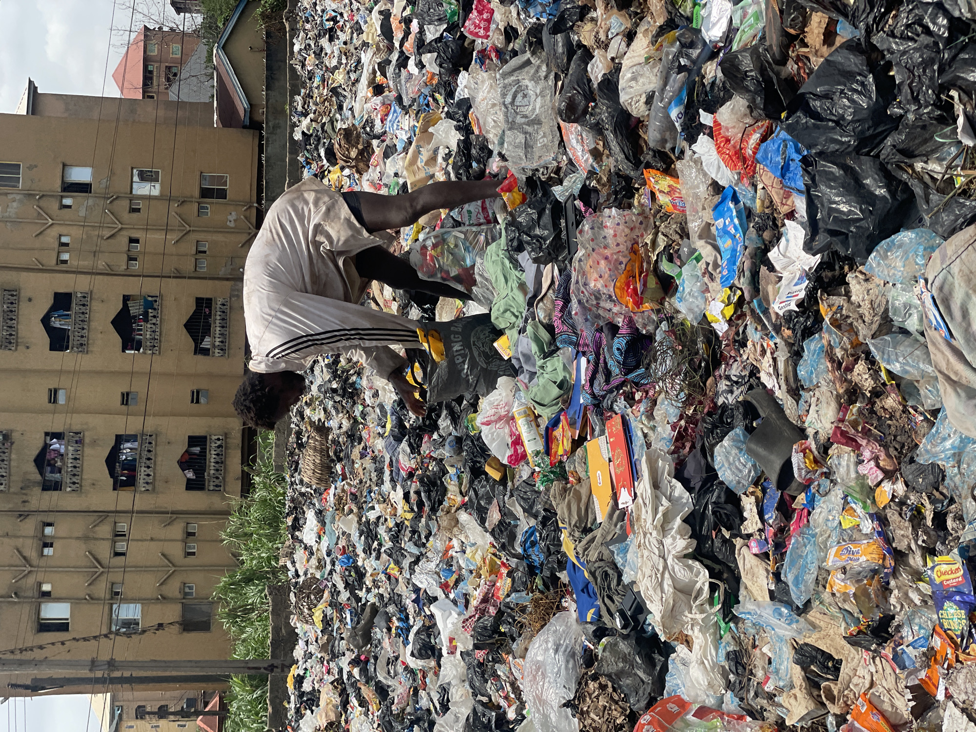 A man is waist-deep in a large pile of garbage. He is wearing light-colored clothing with dark stripes on the sides. He is sorting through the trash, picking out items, and placing them into a bag. The garbage pile is composed of various materials, including plastic bags, food wrappers, and other discarded items. In the background, there is a multi-story building with windows and balconies. The image was taken in Onitsha, Nigeria.