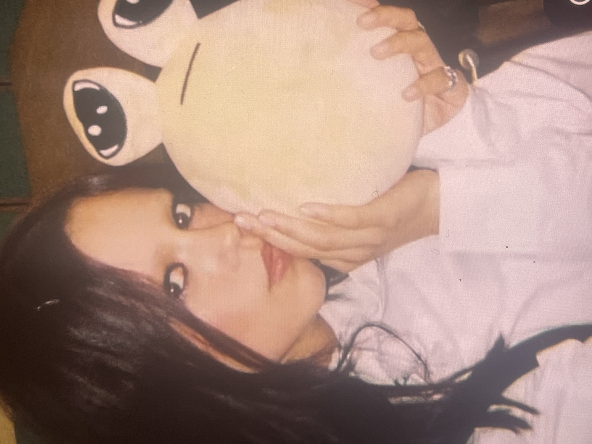 The image captures a close-up of a young woman with long, dark hair, lying down and looking directly at the camera. Her expression is gentle and composed, with dark eyes, defined brows, and lips accentuated with a reddish-pink hue. She appears to be wearing a light-colored, possibly white, long-sleeved shirt.
She is holding a pale yellow, round plush toy with prominent cartoon-like eyes and a simple black line for a mouth. The toy resembles the "Dropout Bear" character. Her hands, adorned with a ring on her right ring finger, are gently cupping the plush against her face, near her nose and mouth, suggesting an intimate or affectionate interaction.
In the background, behind the woman's head and the smaller plush, a larger, dark brown object with similar stylized eyes is partially visible, indicating another plush item. The background is dark and indistinct, implying an indoor setting with low lighting, possibly a bedroom or living space.
The image has a soft, somewhat grainy aesthetic, characteristic of older film photography or a specific digital filter. No specific time of day or weather conditions can be determined. There are no explicit location cues within the image itself to confirm the external context of Poltava, Ukraine. No text is visible. The scene conveys a sense of personal comfort, warmth, and quiet engagement.