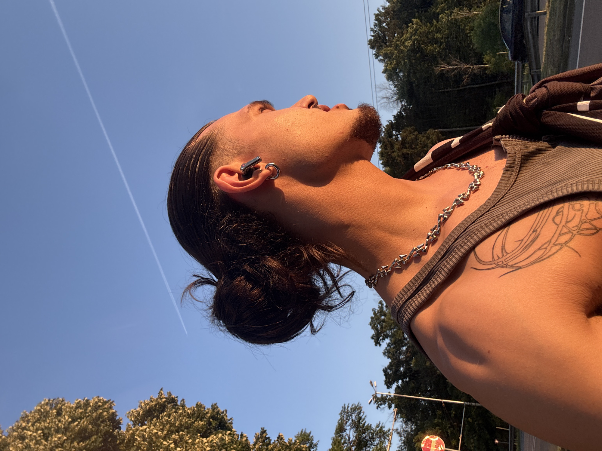 An over-the-shoulder shot captures a young person with dark hair pulled back in a bun, viewed in profile against a bright blue sky. The setting sun casts a warm, golden light on their skin, highlighting their facial features, including a goatee and a large hoop earring. They are wearing a ribbed grey tank top and a chunky silver chain necklace. A tattoo is visible on their left shoulder blade. In the background, trees line a road with a white line and a guardrail. A distant, dark-colored car is parked on the side of the road. The image appears to have been taken outdoors in a park or on the outskirts of a city, potentially Sofia, Bulgaria, given the presence of streetlights in the distance and the general urban-greenery landscape. A contrail streaks across the sky.