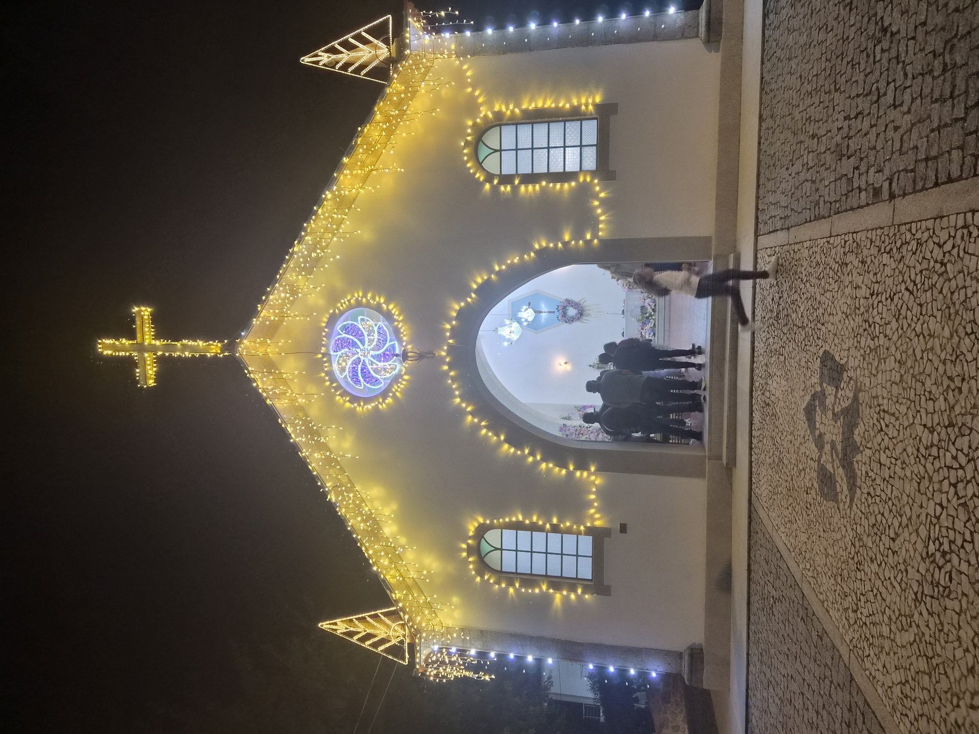 This is a night-time, eye-level, full shot of a small, white chapel in Barcelos, Portugal, elaborately decorated with strings of yellow and white lights. The chapel has a pitched roof topped with a large, illuminated cross. On either side of the roof's peak are triangular structures also outlined in lights. The facade of the chapel is adorned with strings of lights, as are two arched windows. Above the entrance arch, there is a circular stained-glass window with a purple and white geometric design. The entrance archway leads into a well-lit interior where several people are standing, and a chandelier is visible. In front of the chapel, a cobblestone plaza is illuminated by the chapel's lights and streetlights. A young person in dark pants and a light-colored top is captured in motion, running across the plaza from right to left, creating a blur due to the motion and low light. The sky is dark, indicating it is night. The ground is paved with contrasting shades of cobblestones arranged in a patterned design.