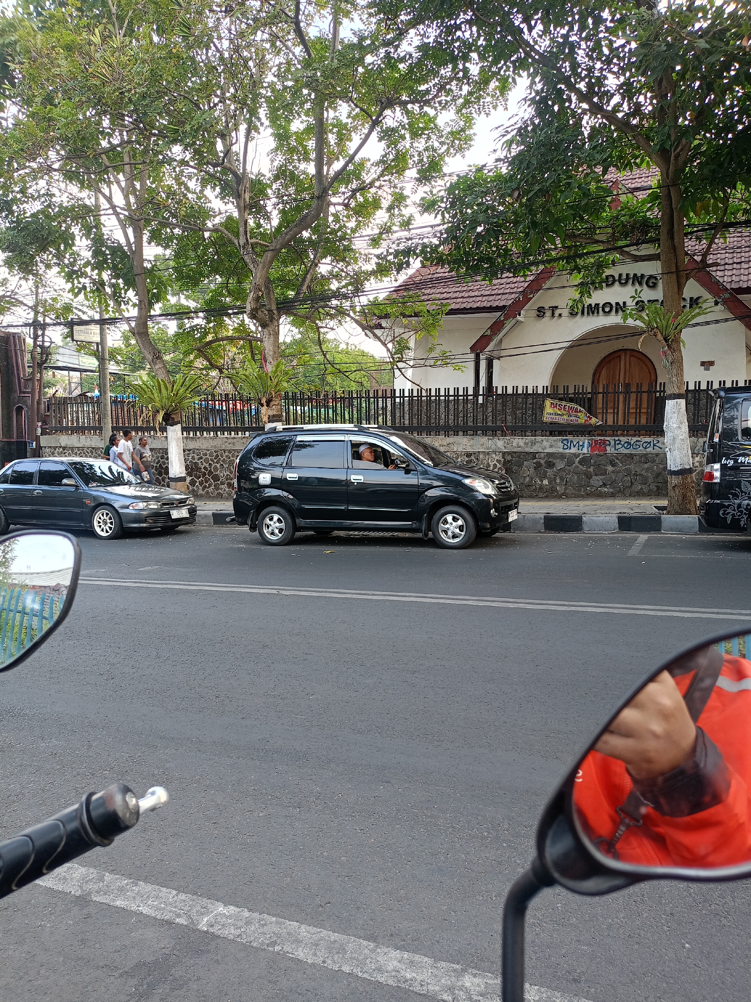 The image shows a street scene in Batu, Indonesia, featuring a church building and vehicles. In the foreground, a motorcycle mirror reflects a person wearing an orange jacket. On the street, a dark gray sedan and a black minivan are parked or stopped. The minivan has a driver visible inside. Behind the vehicles, a stone wall separates the street from a building with a tiled roof. A sign on the building reads "ST. SIMON STOCK." Several trees with green foliage are present, lining the street and partially obscuring the church. The sky is overcast. The road surface is asphalt with white lane markings.