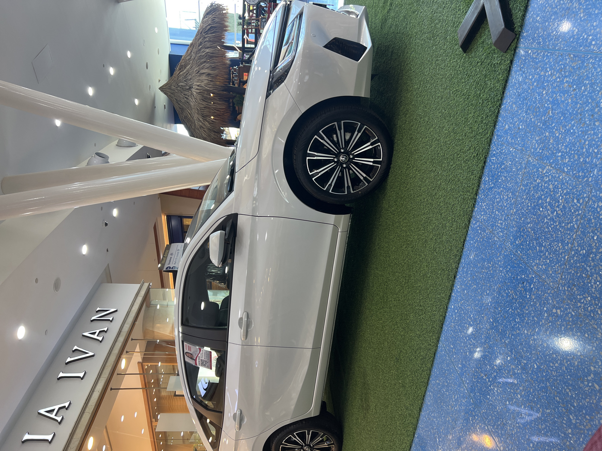 The image displays a white sedan, likely a new model, situated indoors on a strip of green artificial grass. The car features black and silver multi-spoke wheels. Adjacent to the grass, a section of blue speckled flooring is visible. The setting appears to be a brightly lit commercial interior, such as a shopping mall or exhibition space.
In the background, several large white cylindrical pillars ascend towards the ceiling. Beyond these pillars, a distinctive thatched roof structure can be seen, adding an unusual design element to the indoor environment. To the left, a retail storefront with prominent silver letters spelling "LA IVAN" is visible. Further in the background, a sign indicates "Nivel Arriba" (Upper Level) and "Escaleras" (Stairs) with directional arrows, confirming a multi-level building structure. A promotional graphic or QR code is visible through the passenger-side window of the car. No people are present, suggesting the car is on display.