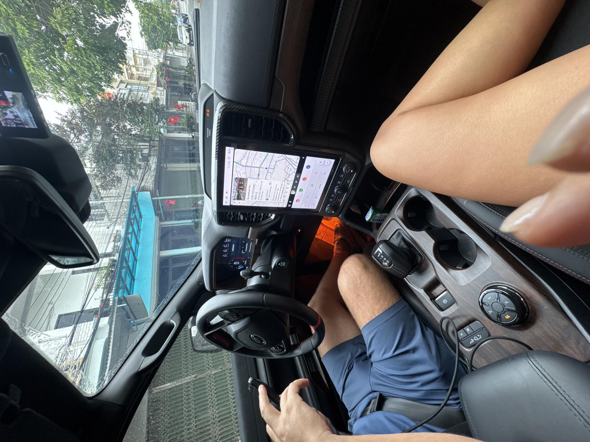 The image shows the interior of a vehicle, specifically the driver's perspective, with a view towards the front. The driver, wearing blue shorts, is visible with one hand on the steering wheel and the other holding a small black object. A female passenger's legs are visible on the center console.
The dashboard features a large infotainment screen displaying a map, indicating a location in Nghia Lo Ward, Vietnam, specifically "CƠM THỐ SUMO" at "10 Điện Biên Nghĩa 14 Lào". The time is shown as 16:54. The gear shift is visible, along with buttons for different drive modes.
Outside the vehicle, buildings and trees are visible, suggesting an urban setting. Vietnamese flags are visible in front of the buildings.