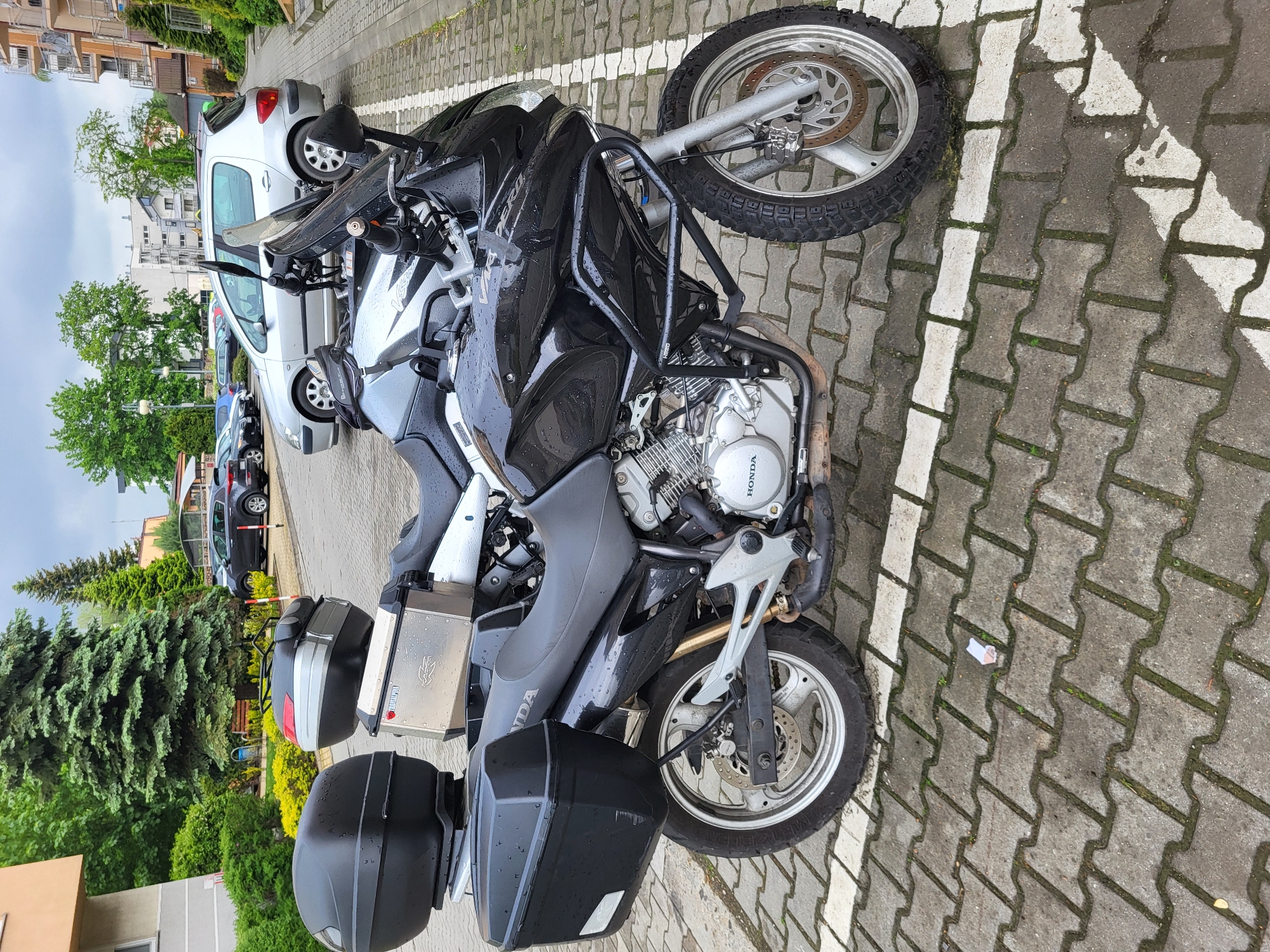 The image depicts a black Honda motorcycle parked on a paved surface with white line markings, indicating a parking space. The motorcycle is equipped with storage boxes on the rear and appears to be wet, suggesting recent rain. In the background, several parked cars are visible, along with some buildings and trees, suggesting an urban or suburban environment. The location is within the city of Unknown, Unknown.