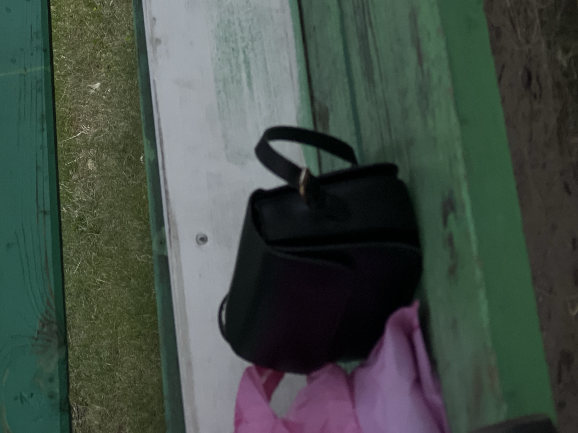 The image shows a black handbag placed on a green wooden bench. Next to the handbag is a crumpled pink object, possibly a bag or piece of clothing. The bench appears to be outdoors, with grass visible in the background. The scene is likely in Ulyanovsk, Russia.