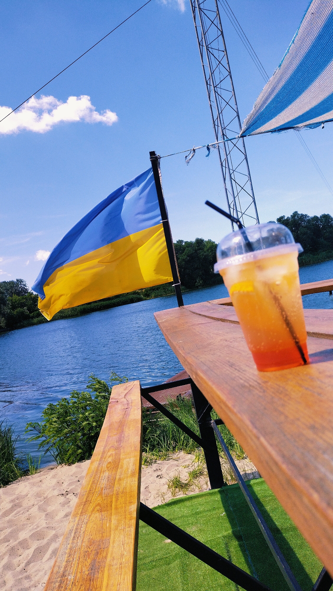 In Horishni Plavni, Ukraine, the image depicts a waterfront picnic table with a drink on it, next to a Ukrainian flag. The wooden table is positioned on a patch of artificial grass, overlooking a sandy bank and a body of water, likely the Dnipro River. A tall metal tower, possibly part of a broadcast or communication structure, rises in the background, partially obscured by a striped blue and white awning. The flag of Ukraine, with its distinct blue and yellow horizontal bands, flutters in the breeze. A clear plastic cup filled with an orange beverage and ice, with a black straw, sits on the table. The sky is a clear blue with a few scattered white clouds. The surrounding vegetation consists of green trees and bushes along the riverbank.