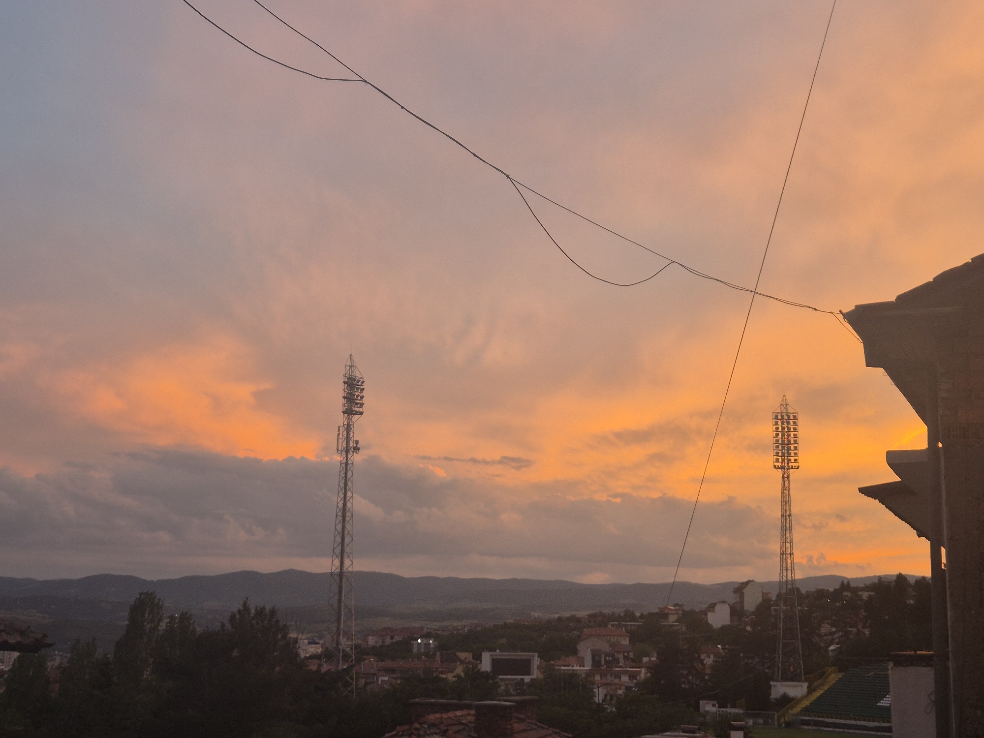 The image captures a wide shot of a city at sunset, likely from an elevated vantage point. The sky is a vibrant gradient of orange and pink hues, with scattered clouds adding texture. Two tall, industrial-looking towers with multiple lights are prominent features in the landscape, one on the left and another on the right side of the image. Below them, a cluster of buildings, characteristic of a residential or commercial area, is visible, nestled amongst trees and rolling hills in the background. A dark, angled structure, possibly a rooftop or a building facade, occupies the right foreground, partially obscuring the view. Power lines are strung across the upper portion of the frame, contributing to the urban setting. The scene conveys a sense of twilight and the transition from day to night.