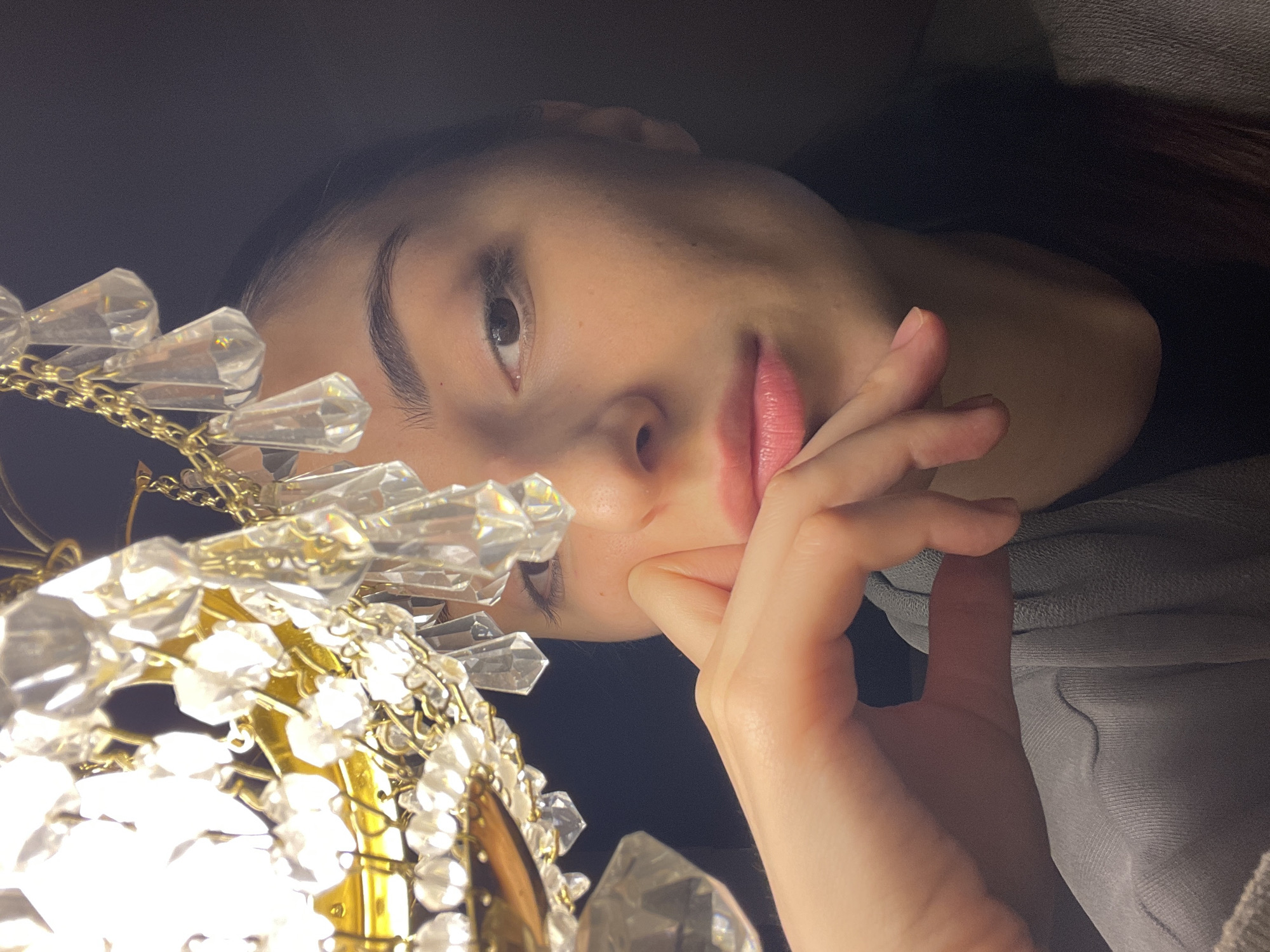 This image presents a close-up portrait of a female individual in an interior location within Moscow, Russia. A prominent, illuminated chandelier, composed of clear crystal pendants and gold-toned metal components, occupies the upper-left foreground, casting a bright glare and partially obstructing the view. The individual is positioned to the right, facing forward. Her right eye, with a brown iris, is open and directed towards the viewer. Her right hand, with unpainted fingernails, is positioned against her lower face, covering the chin and part of the cheek. Her lips are a light pink. She wears a light gray, possibly ribbed, long-sleeved garment. The background is dark and featureless, consistent with dim ambient lighting aside from the chandelier.