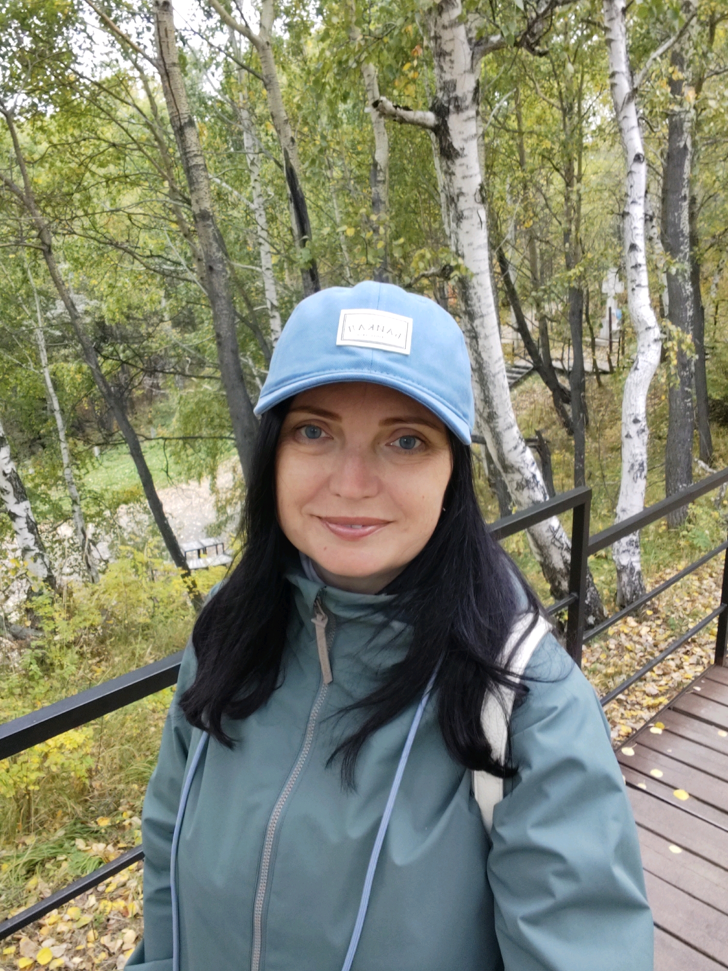 A woman with dark hair and blue eyes, wearing a light blue baseball cap and a teal jacket, smiles at the camera. She is standing outdoors on a wooden pathway next to a black metal railing, with a backdrop of birch trees and foliage. The setting appears to be a park or natural area, possibly within the city limits of Nikola, Russia, given the birch trees common to the region. The presence of fallen leaves suggests it is autumn.