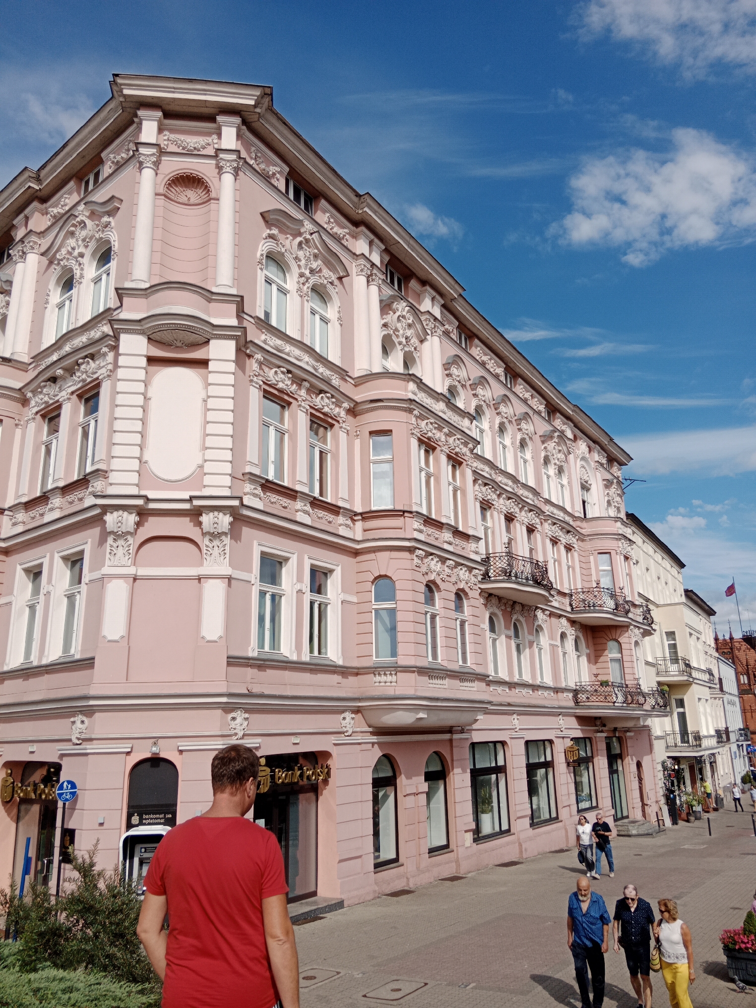 The image captures a bright, sunny day in Bydgoszcz, Poland, focusing on a distinct pink building with ornate architectural details. The building, with its multiple stories, arched windows, and decorative balconies, appears to be a historic structure, likely housing commercial establishments on the ground floor. The presence of a "Bank Polski" sign on one of the storefronts confirms it's a bank.
In the foreground, a man wearing a red t-shirt is walking away from the camera, his back towards the viewer. He is walking on a paved sidewalk adjacent to the building. Further down the street, a few other individuals are seen walking, including a couple in the middle distance and two men and a woman walking towards the camera in the lower right quadrant. The street is paved with cobblestones and lined with more buildings and some greenery, suggesting a well-trafficked urban area. The sky above is a vibrant blue with scattered white clouds. The overall scene depicts a typical day in a European city, with people going about their business in front of charming historical architecture.