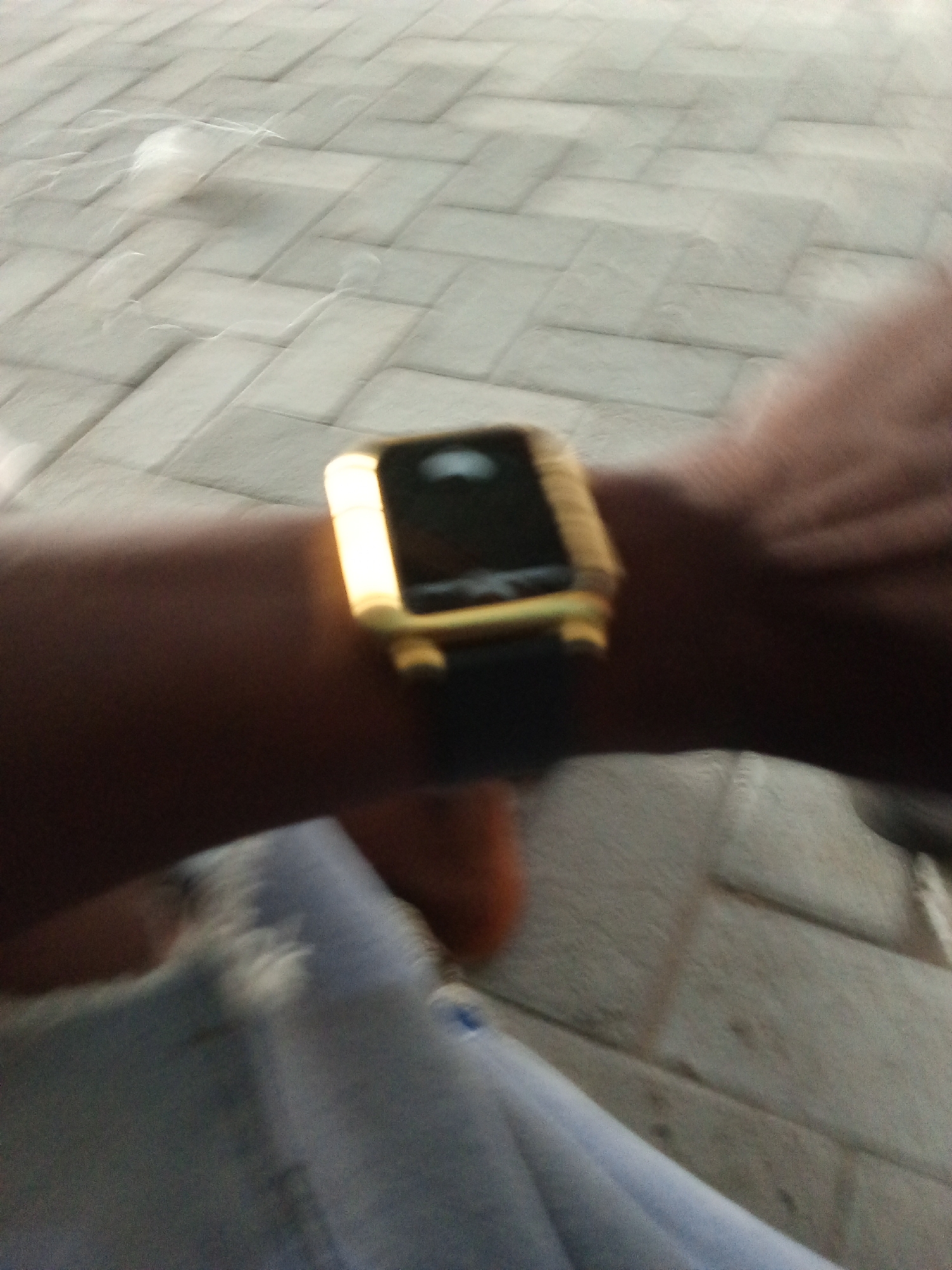 A close-up, blurry shot shows a person's arm and wrist wearing a gold-colored smartwatch with a black strap. The smartwatch face is dark and reflective, with a bright light reflecting from the top right. The person's arm is dark-skinned. The background is a blurred, grey, brick-like pattern on the ground. A portion of light-blue clothing is visible in the lower left of the frame. The scene appears to be outdoors.