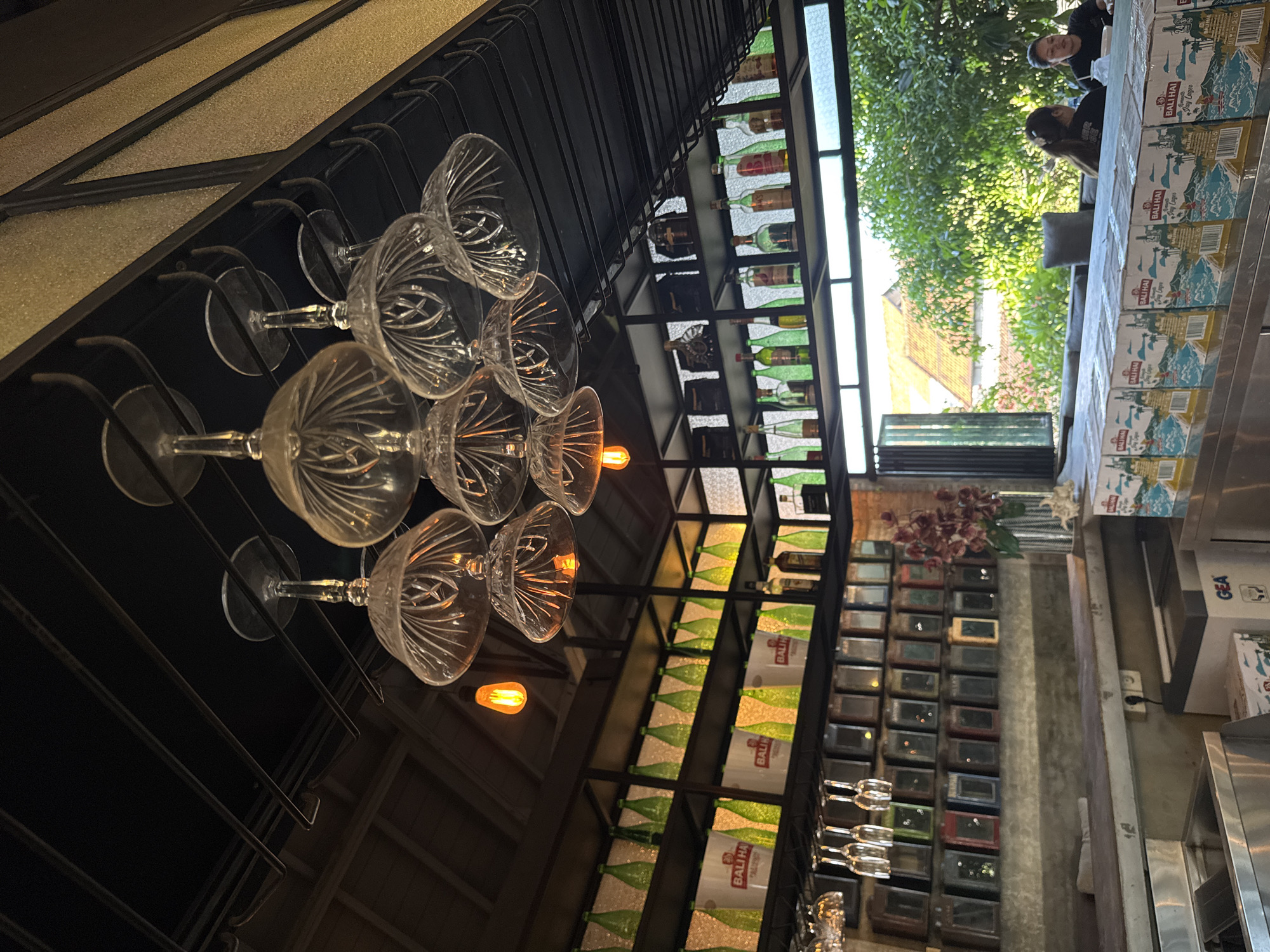 In North Kuta, Indonesia, this indoor-outdoor bar features a shelf filled with various green bottles and white buckets displaying "BALI HAI" branding. Above the shelf, crystal wine glasses are hung upside down, illuminated by a vintage-style orange lightbulb. The bar area is visible in the foreground, with stainless steel surfaces and a counter. Through an open window, a lush green outdoor seating area with trees and buildings with tiled roofs can be seen. Two women are seated at the outdoor bar, one facing the camera and the other with her back to it. The scene is brightly lit, with a focus on the bar's decorative elements and the inviting outdoor ambiance.