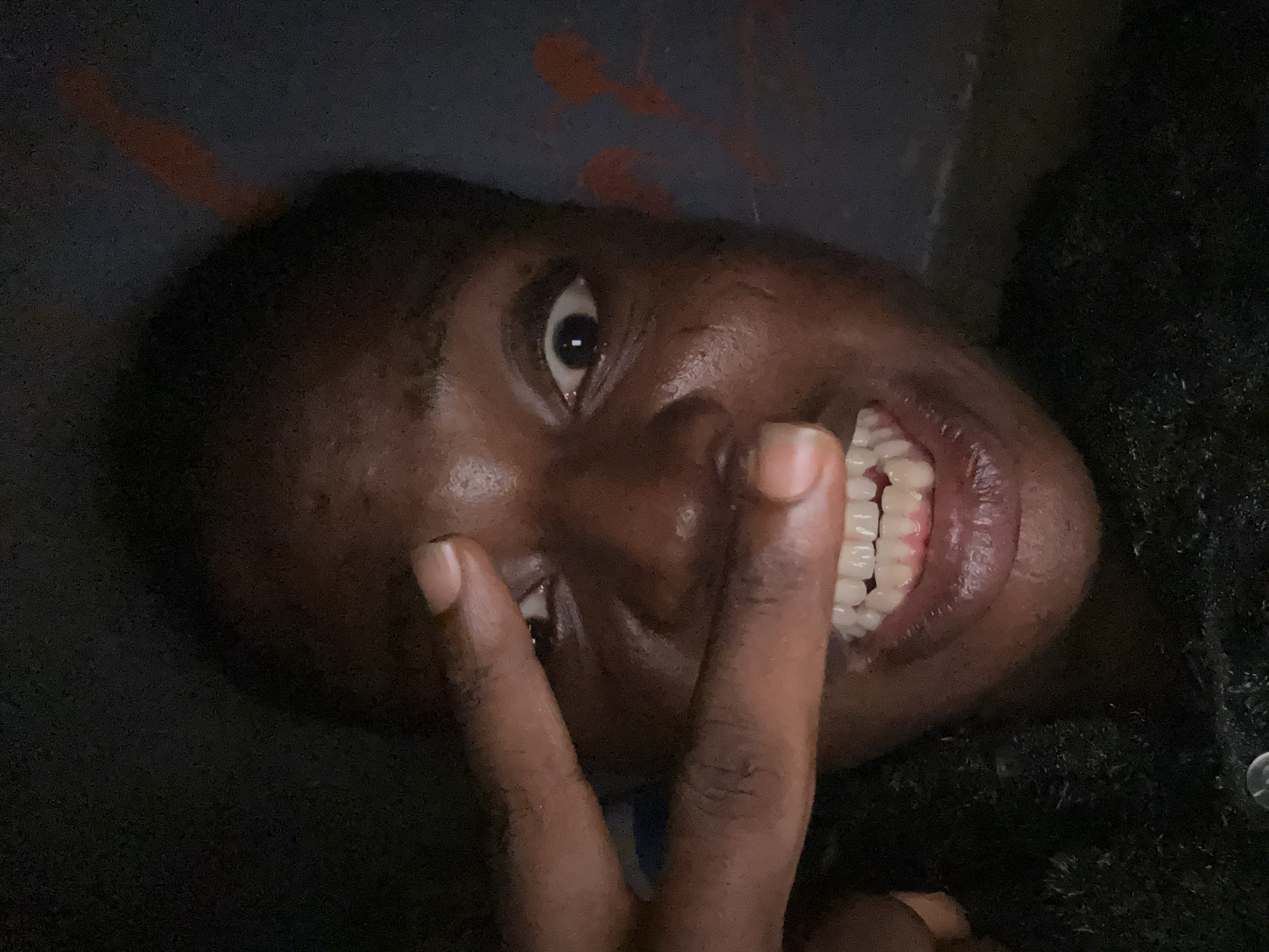 A close-up, angled shot shows a person with dark skin smiling widely while making a peace sign with their hand. The background is dark and indistinct, with a few vague orange marks. The individual's teeth are visible, and the focus is mainly on their face and hand. The location is not specified.
