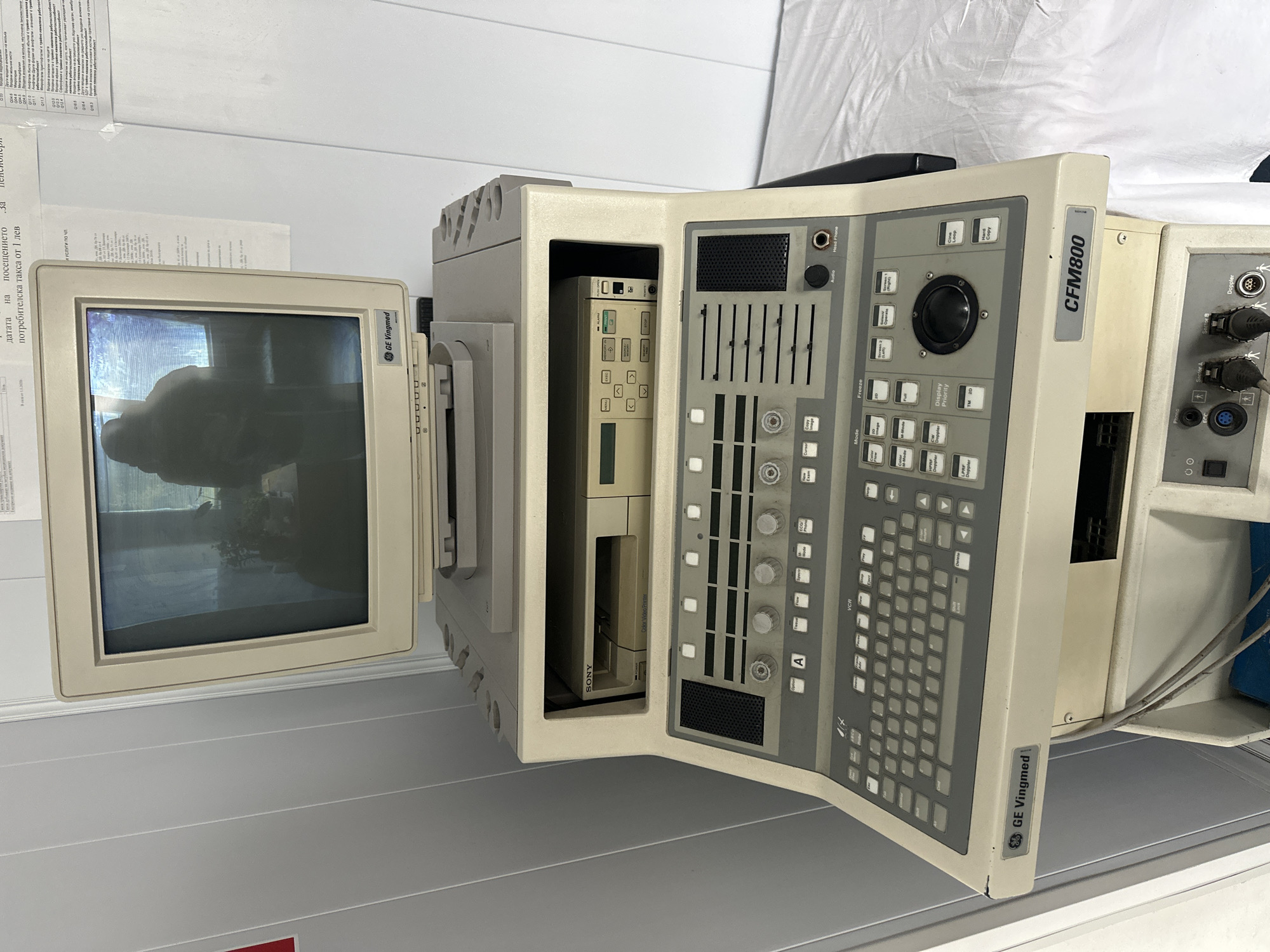 The image shows a vintage GE Vingmed CFM800 ultrasound machine. The machine consists of a CRT monitor, a central processing unit, and a control panel. The monitor is off, displaying a reflection of the surrounding environment. The CPU has a Sony color video printer and other internal components visible. The control panel is open, revealing a keyboard, sliders, knobs, and buttons. Cables are visible connecting parts of the system. The ultrasound machine is in a medical facility. A document with Bulgarian Cyrillic text is partially visible, referencing a "потребителска такса от 1 лев" (consumer tax of 1 lev) and text that contains a reference to "работоспособност" (work capacity) and "Zemen, Bulgaria". The surfaces surrounding the machine appear to be part of a medical room setting.