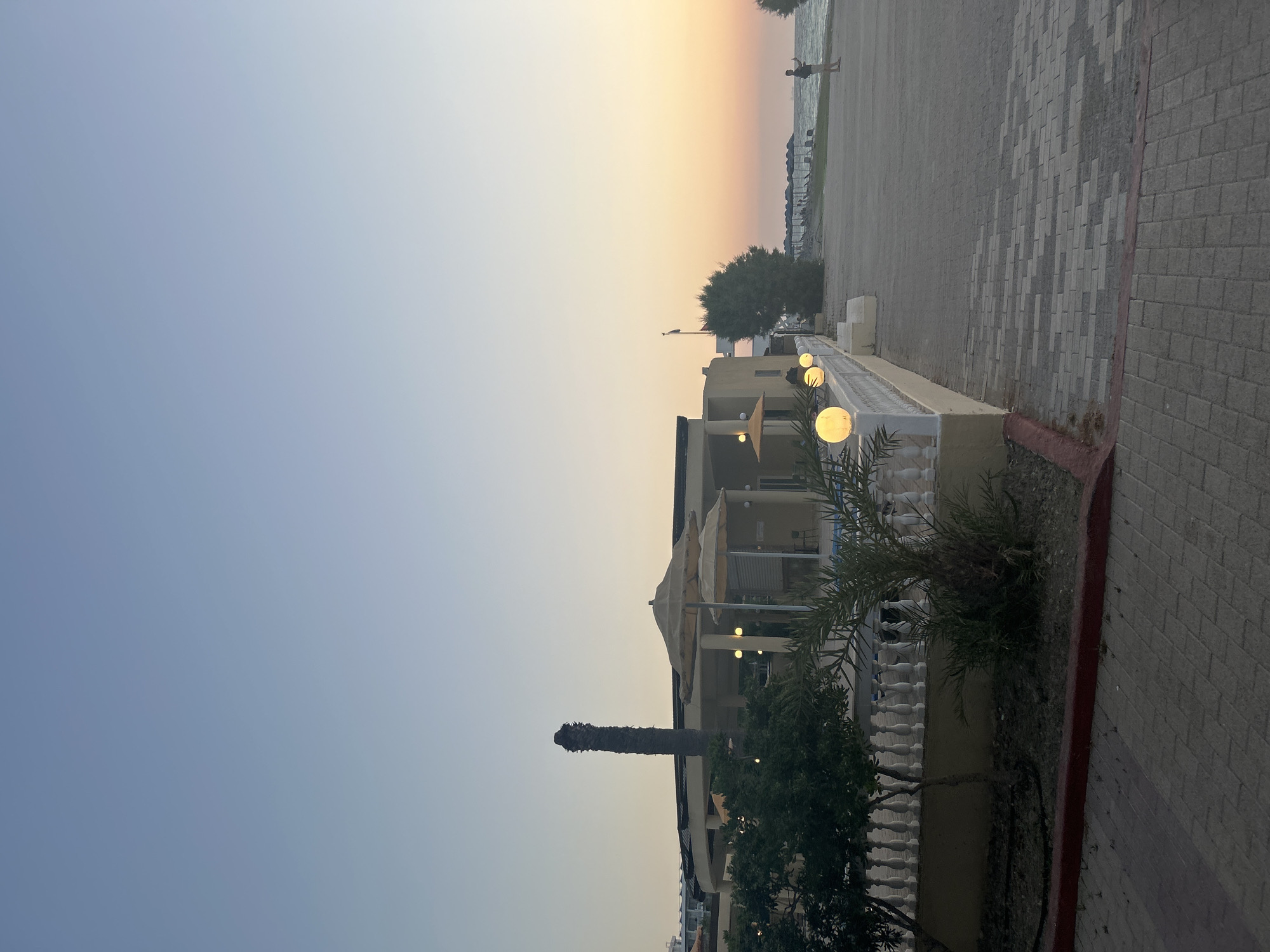 The image depicts a scenic view of a coastline at dusk, likely in Rhodes, Greece. The sky transitions from a pale blue near the top to a warm gradient of orange and yellow near the horizon, indicating sunset. A body of water, possibly the sea, is visible in the background.
In the mid-ground, a building with a light-colored facade and multiple windows stands beside a paved walkway. Outdoor lights, spherical and emitting a warm glow, are mounted along a white decorative railing in front of the building, suggesting an evening ambiance. Trees and palm fronds add greenery to the scene, with one notable palm tree trunk appearing stark and textured.
On the paved area near the water's edge, a solitary figure is visible, appearing to walk towards the sea. The walkway is made of interlocking paving stones in a patterned arrangement. The overall setting evokes a relaxed atmosphere, common in coastal resorts. The time of day is clearly evening, and the weather appears clear. There is no visible text in the image.