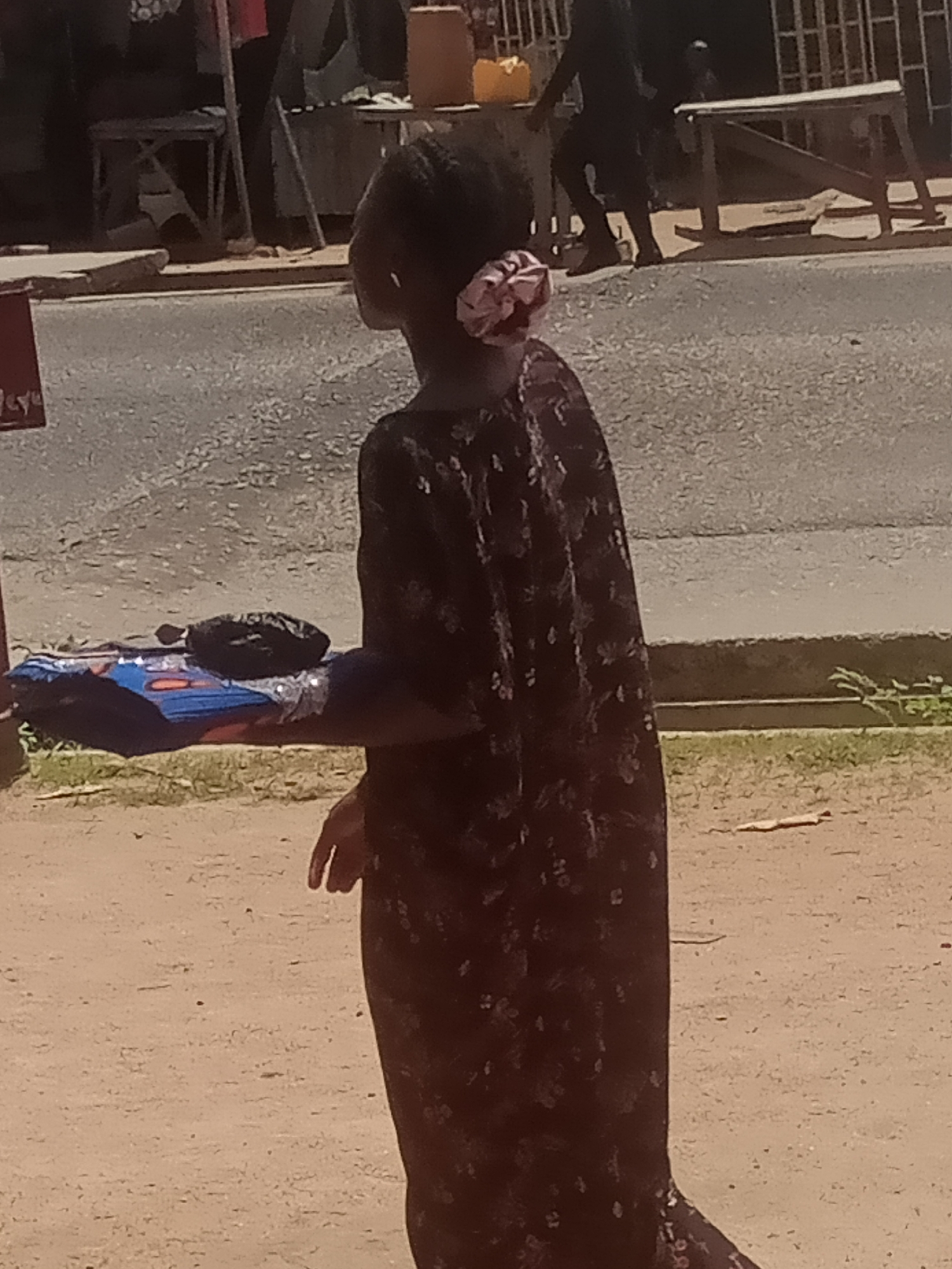 A young girl with a pink and white hair accessory stands facing away from the camera. She is wearing a dark-colored dress with a floral pattern. To her left, on the ground, is a rolled-up mat or blanket, dark blue with some red and white patterns, and a dark object on top of it. The girl is standing on a dirt surface, with a paved road behind her. In the background, there are several structures that appear to be market stalls or temporary shelters, with people visible in the distance. The overall setting suggests a market or street scene in a city. This image appears to have been taken in Jalingo, Nigeria, judging by the context.