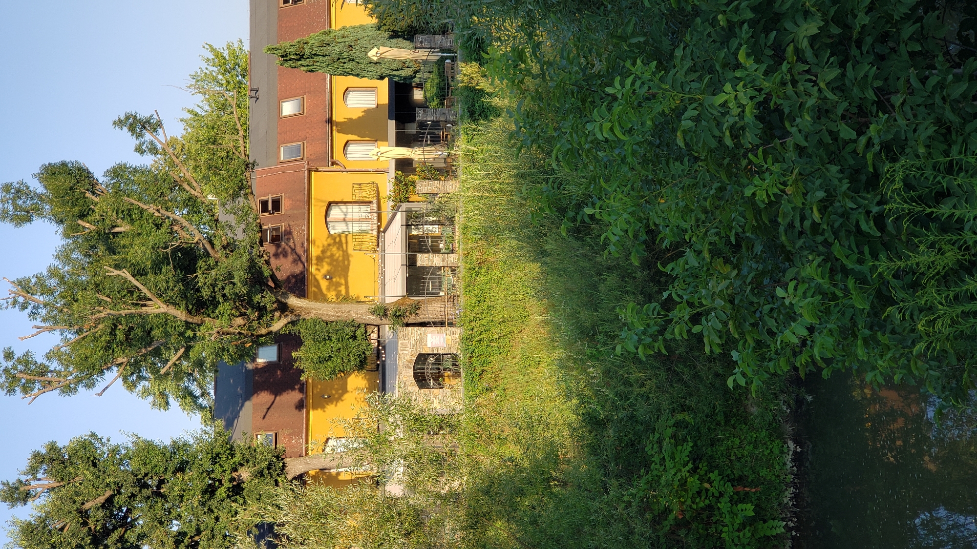 The image shows a building with a yellow facade and a brown roof, situated next to a body of water. Trees with green foliage are in the foreground and surrounding the building. Some of the trees appear to be mature, with thick trunks and branches. The building has multiple windows and doors, and there are outdoor seating areas with umbrellas. The overall scene suggests a peaceful, possibly tourist-oriented location. The location is identified as Prešov, Slovakia.