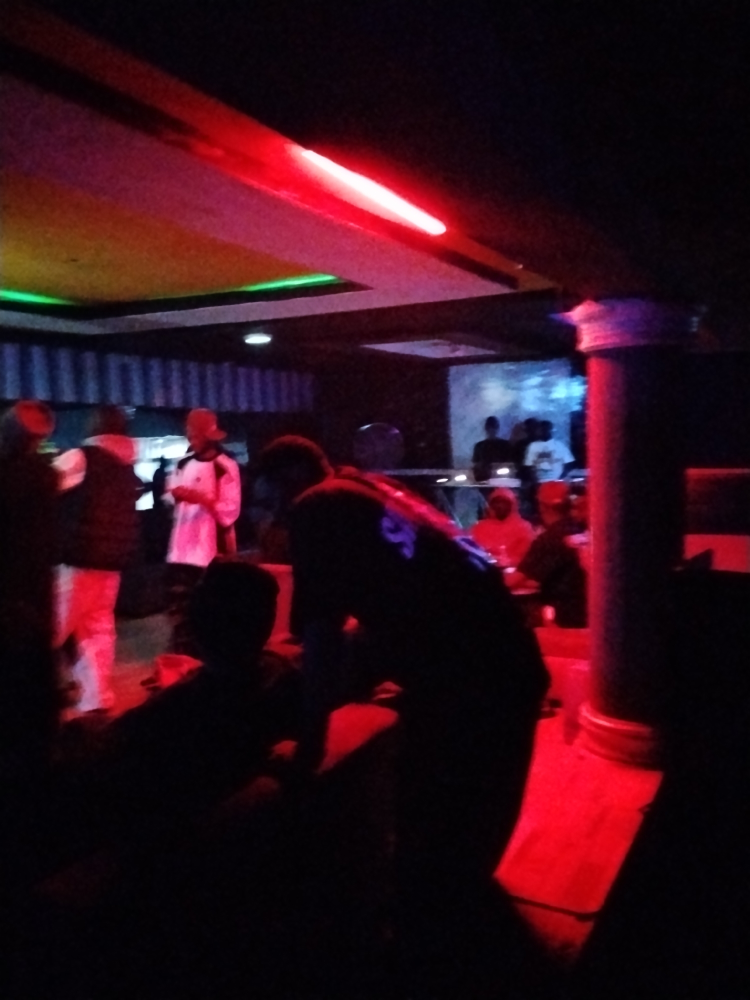 The image captures an indoor scene, possibly a club or lounge, characterized by low lighting and vibrant neon colors. A prominent red neon light streaks across the ceiling, casting a reddish hue on the surroundings. Green neon lighting is visible along a recessed ceiling panel. In the foreground, a group of people are silhouetted against the dim light, with some figures more distinct than others. One individual in the mid-ground appears to be wearing a white long-sleeved shirt and a cap. Another person in the mid-ground is wearing a white puffer vest over a white garment and light-colored pants. Further back, a DJ setup is partially visible with individuals standing behind it. The environment appears to be a public gathering space. The image does not provide specific details to identify the location within Durumi, Nigeria.
