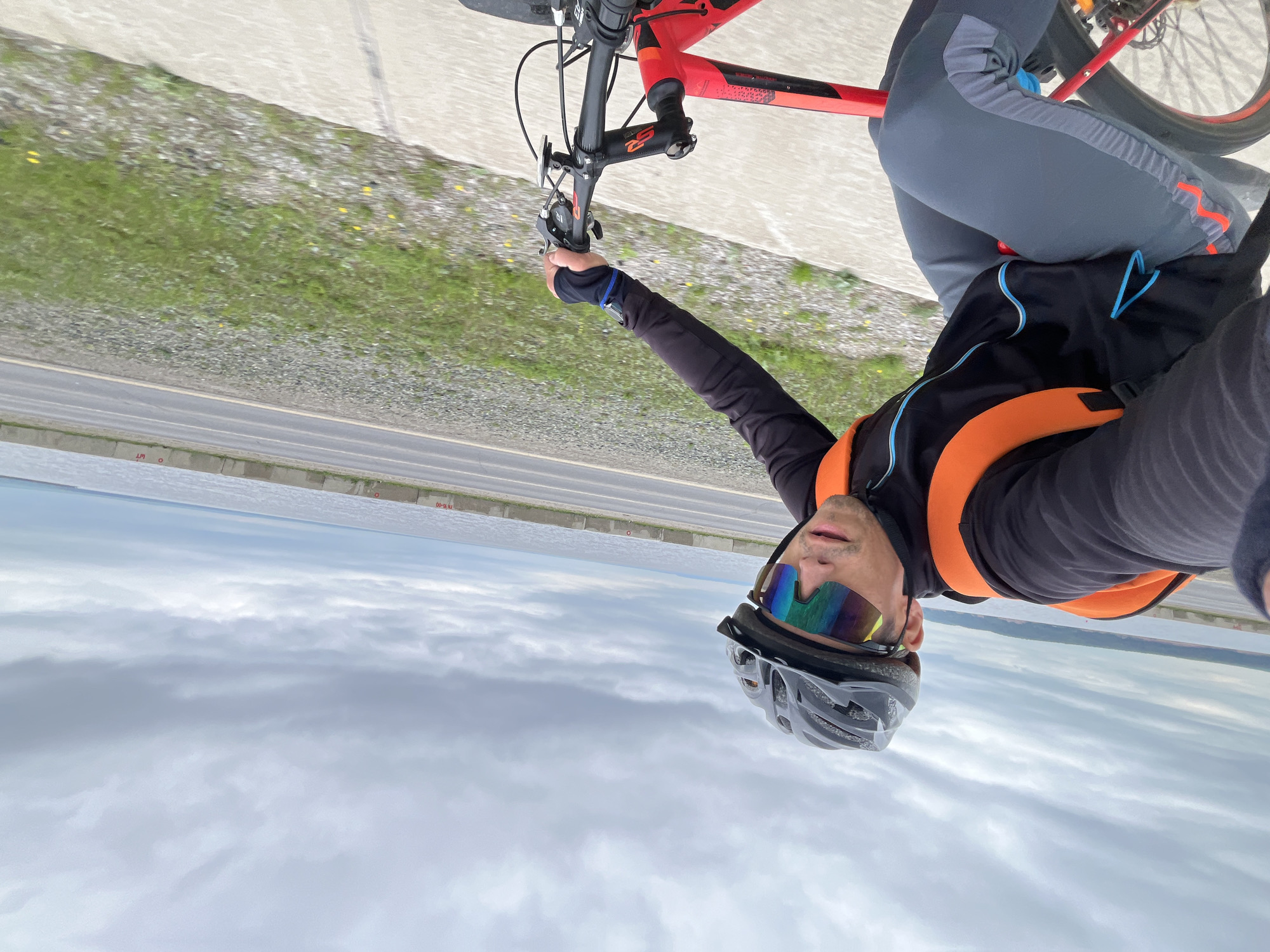 An extreme wide-angle, POV shot captures a person cycling on a paved path next to a road, with a body of water in the background. The cyclist wears a black helmet, a black long-sleeved jacket with bright orange straps for a backpack, and gray cycling pants with orange and blue accents. They also wear mirrored cycling glasses. The cyclist's left hand grips the handlebars of a red bicycle. The environment is overcast, with a cloudy sky. The road is adjacent to a concrete barrier separating it from a strip of green grass. The water is calm, reflecting the overcast sky. The scene appears to be along the Kama River embankment in Chaikovsky, Russia, a location known for its scenic river views and recreational paths.