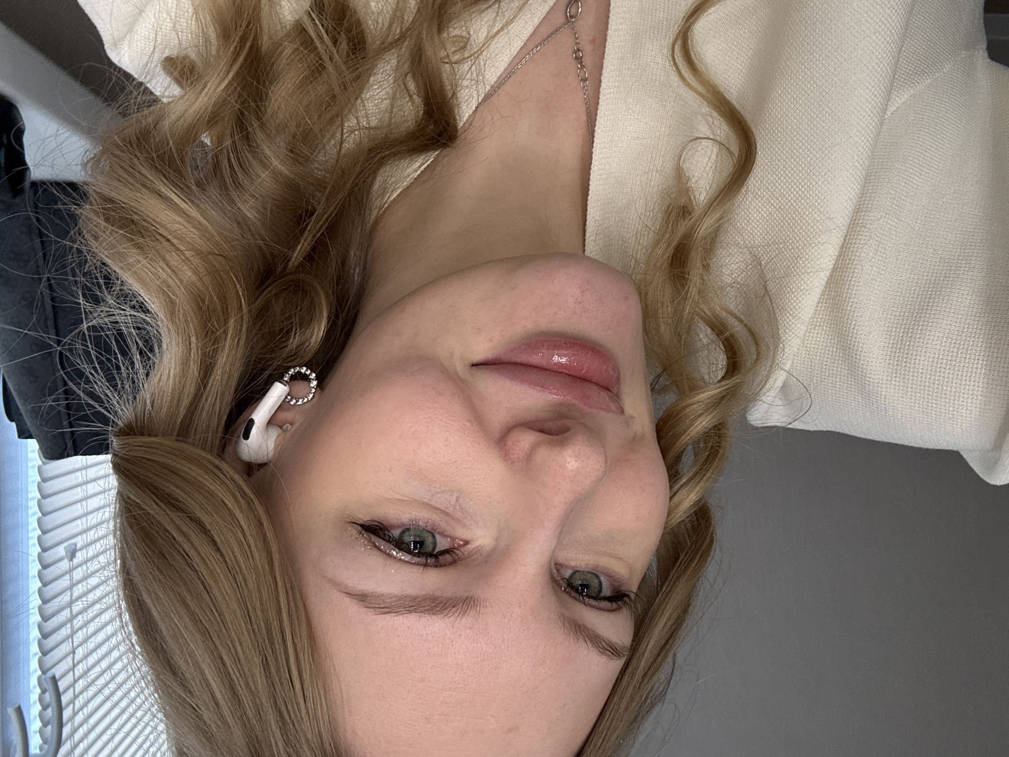 A close-up, angled selfie of a light-skinned woman with light brown, wavy hair and gray-green eyes. She is wearing an off-white textured top. Her lips are glossy and her eyes are lined with dark eyeliner. An Apple AirPods earpiece adorned with a jeweled ring is visible in her left ear. A delicate silver necklace is visible at her neck. The background is indistinct, showing a partially visible window with white blinds on the left, and a plain gray wall on the right. The location is not ascertainable from this image and there is no indication that it is located in Perm, Russia.