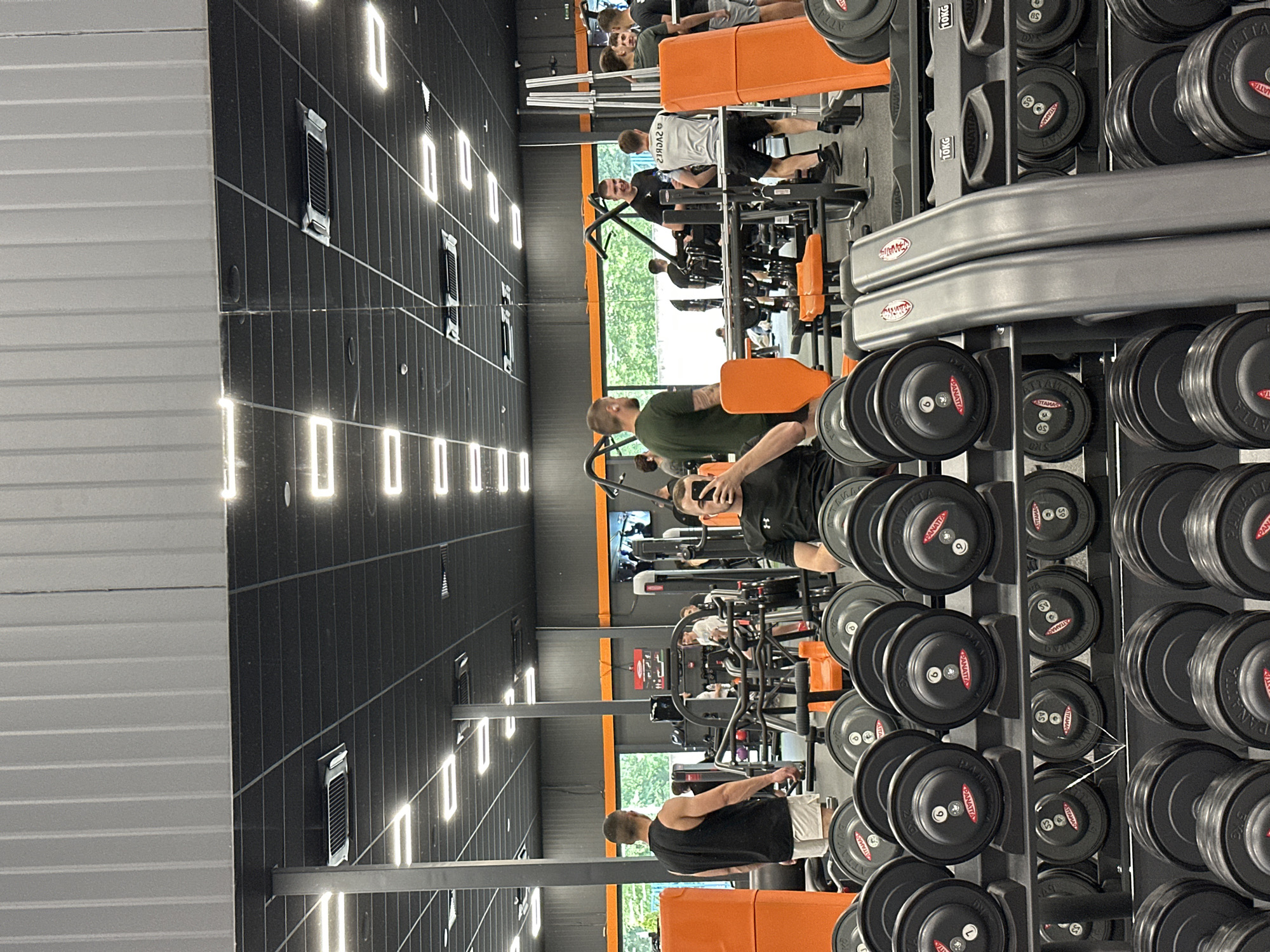 The image captures the interior of a gym in Plovdiv, Bulgaria. The space is well-lit with a black ceiling adorned with rectangular light fixtures. A rack of dumbbells dominates the right side of the frame, showcasing a range of weights labeled with the brand "PANATTA". Various gym-goers are visible, engaging in exercises on weight machines. The machines are predominantly orange and black in color. Through the gym's windows, a green landscape is faintly visible, suggesting an outdoor view. Ventilation ducts can be observed along the ceiling.