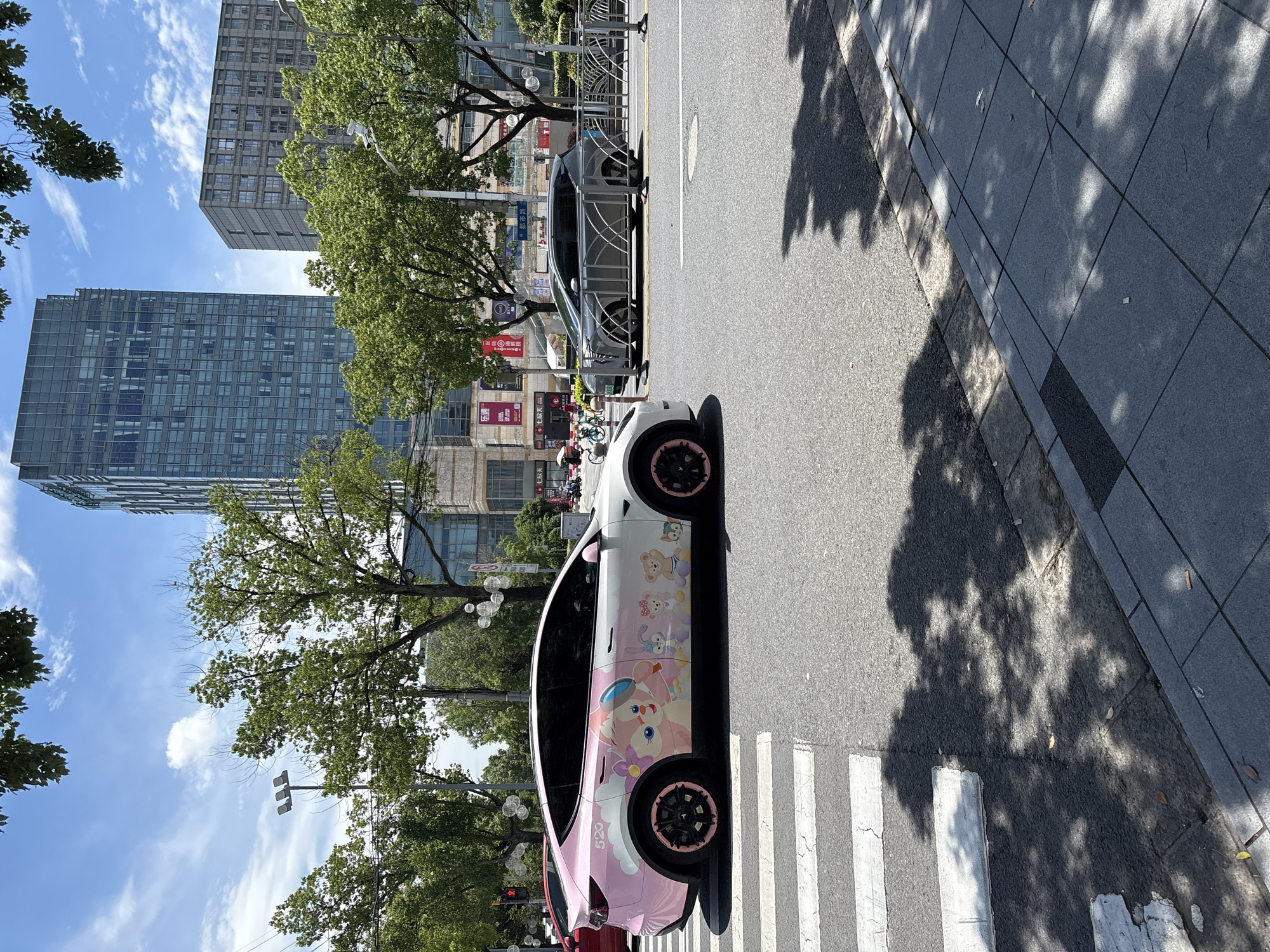 In Minhang District, China, a white Tesla Model Y with a pink cartoon decal wrap featuring characters like StellaLou and CookieAnn is parked on a street. The car has pink accents on its black rims. To the right of the Tesla, a gray minivan is driving on the road. In the background, a tall glass skyscraper and a concrete building stand amidst lush green trees. Pedestrians and bicycles are visible on the sidewalk, and a crosswalk is present in the foreground. The sky is blue with scattered clouds, and sunlight casts shadows on the ground.