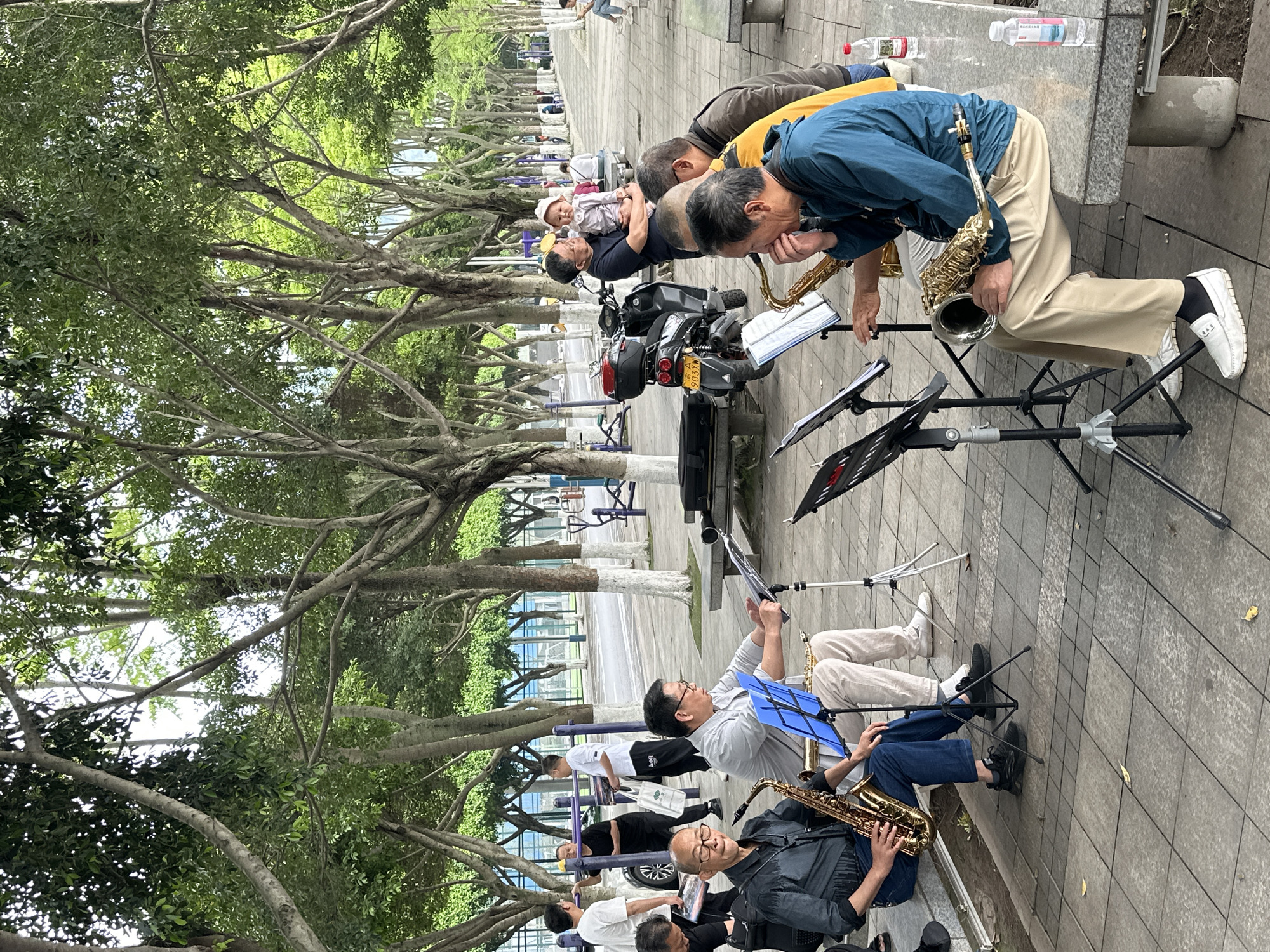 The image depicts a group of people playing saxophones in an outdoor setting, possibly a park or public space. The location is likely Jiangjin, China. Several individuals are positioned along a paved walkway, seated or kneeling with music stands in front of them, playing their saxophones. Some are seated on benches. They are of varying ages. There is a motorcycle parked among them. The area is shaded by trees with intertwined branches, casting shadows on the walkway. In the background, there are more trees and some structures. Bottles of water are visible near the group.