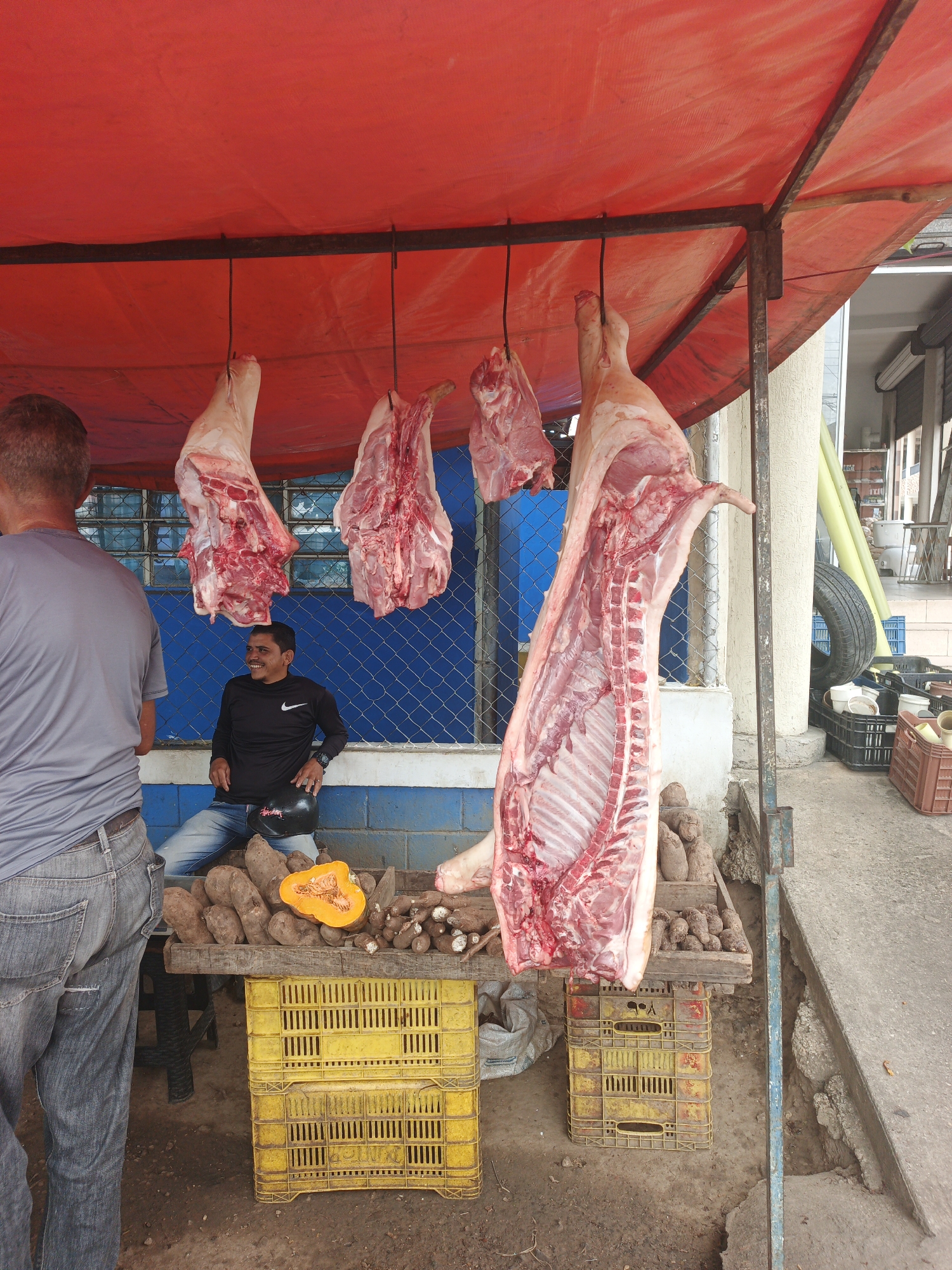 A daytime view in Tinaquillo, Venezuela, shows a rudimentary outdoor market stall with a red tarp roof. Hanging from the stall's frame are four cuts of pork, including a full side of pig with visible ribs. Below the meat, the stall displays yams and half of a cut pumpkin on a wooden surface supported by stacks of yellow plastic crates. A man in a black long-sleeved shirt and jeans sits behind the stall, smiling. A second man, wearing a grey t-shirt and jeans, stands to the left, facing the stall. In the background, behind the stall's display, is a blue wire fence. To the right of the stall, there are stacks of plastic crates and a spare tire.
