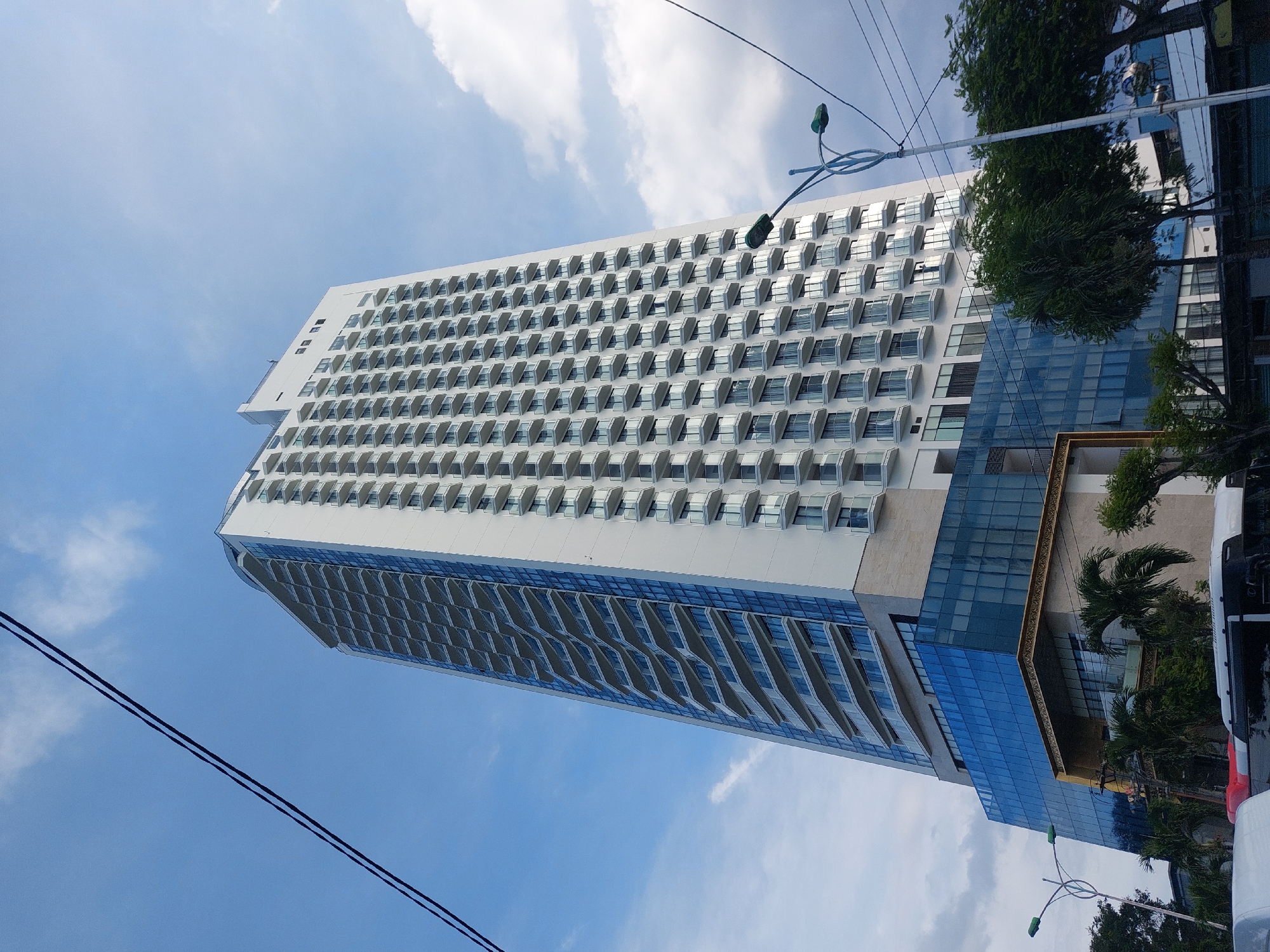 This is a low-angle, eye-level shot of a modern, white high-rise building with many balconies. The building is located in Nha Trang Ward, Vietnam. The building has a glass facade on the lower levels, which reflects the blue sky and white clouds. There are power lines in the foreground, and some trees and other buildings can be seen at the base of the high-rise. The sky is a clear blue with a few scattered white clouds.