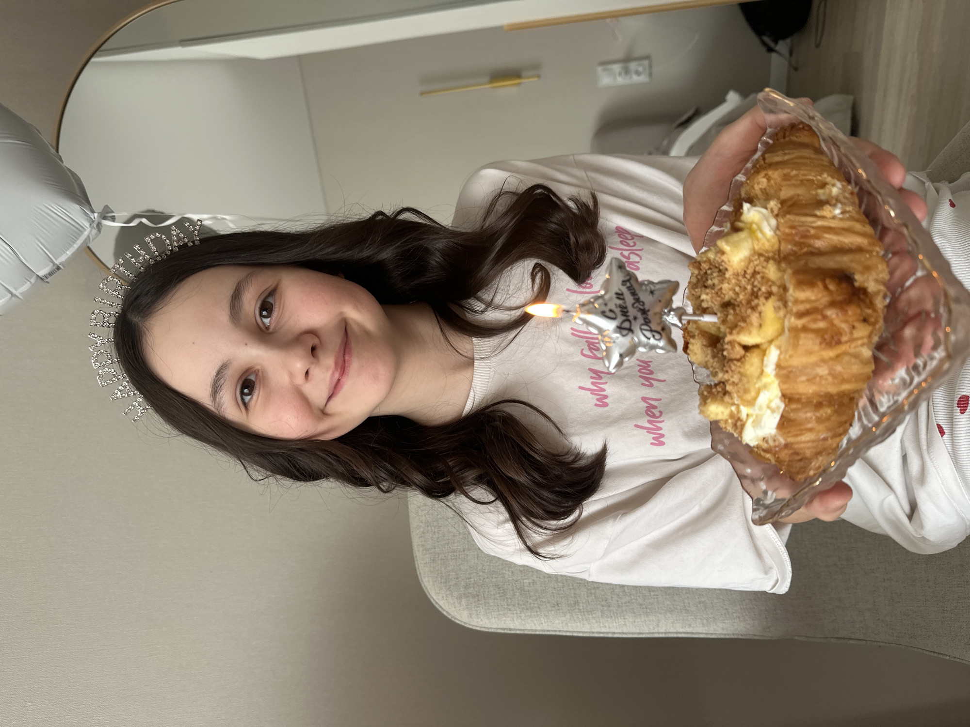 A young person, likely a female, is seated indoors, holding a croissant in a clear container. A star-shaped candle is stuck into the croissant, with a flame visible. The individual is wearing a "Birthday" tiara and a t-shirt with pink writing. The background is a simple interior setting with off-white walls and a glimpse of a balloon and a mirror. There's no explicit information in the image connecting it to Votkinsk, Russia.