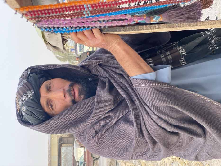 The image depicts a street scene in Kandahar, Afghanistan, centered on a middle-aged to elderly man standing outdoors. The man, with weathered skin, dark eyes, and a short dark beard, wears a dark grey or black turban with a patterned accent and a dark grey or brown shawl draped over his head and shoulders. A light blue long-sleeved garment is visible beneath the shawl. His right hand grips a rustic wooden display stand from which numerous strands of brightly colored beads are suspended. These beads, likely prayer beads or necklaces, appear in various hues including red, orange, pink, light blue, dark blue, black, and purple, some featuring tassels.
In the blurred background, a street or market environment is discernible. On the left, a portion of a red truck or cart is visible next to an industrial-style building. Further back, at least two other male figures, also wrapped in shawls, are present. A light-colored vehicle is partially obscured in the mid-ground. The ground appears unpaved or dusty. The overall lighting is diffused, suggesting an overcast sky.