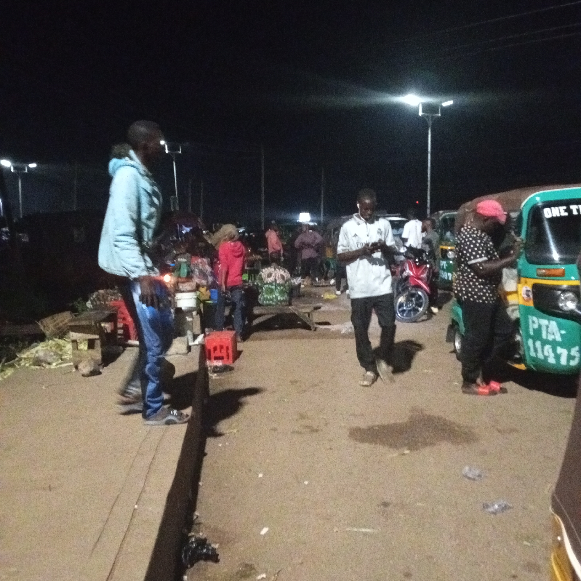 The image depicts a nighttime scene at a market in Bukuru, Nigeria. Several individuals are present, some standing and others walking. In the foreground, a person in a light blue jacket and blue jeans is walking away from the camera. To the right, another individual in a white t-shirt and black pants is looking at their phone. Further to the right, a person wearing a red cap and a polka-dot shirt is also looking at their phone, standing next to a green and yellow motorized tricycle. The tricycle has the text "ONE TI" visible on its side and a license plate that reads "PTA-11475". In the background, other people and market stalls are visible under streetlights. The ground is paved and appears to be a street or market area. There is some debris scattered on the ground.