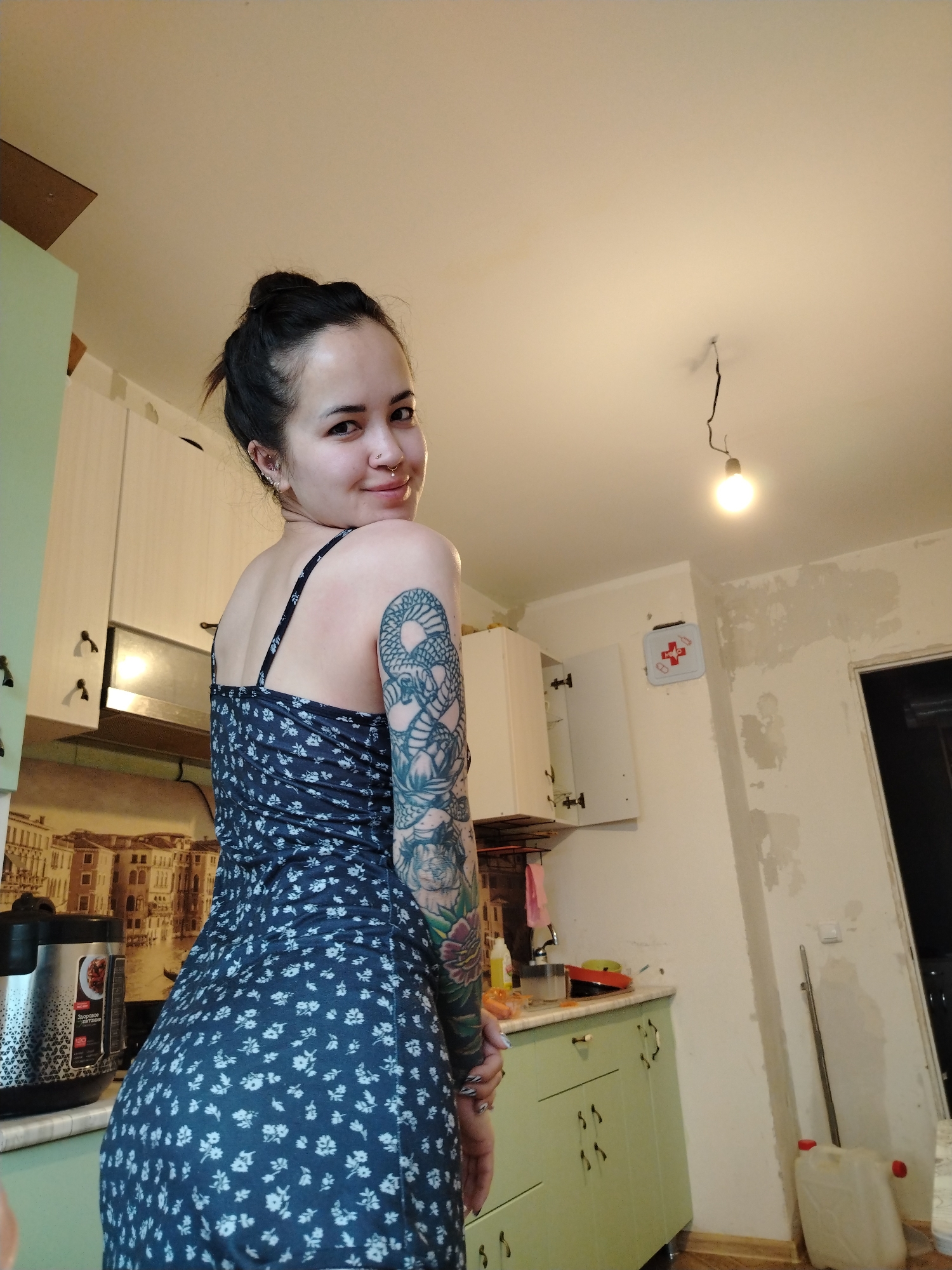 A woman with a tattoo on her left arm is standing in a kitchen. She is wearing a dark blue dress with white floral patterns. Her hair is pulled back in a bun. She is looking over her shoulder and smiling. The kitchen has light green and white cabinets. A slow cooker is visible on the countertop. There is a first aid kit on the wall. The wall is partially plastered with unpainted patches. A bare light bulb hangs from the ceiling. The location appears to be Noviy, Russia.