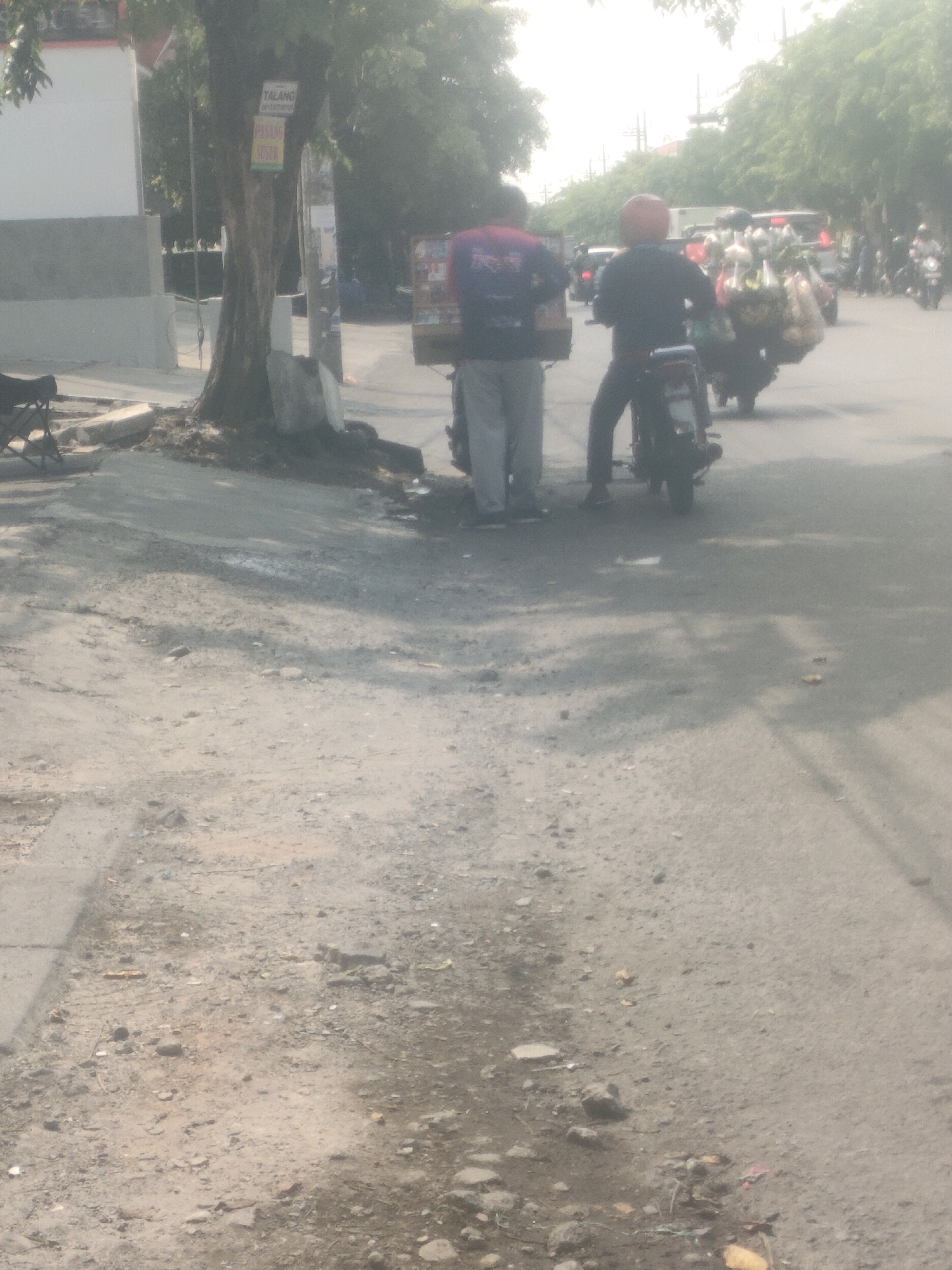 The image captures a street scene in Surabaya, Indonesia, during daylight hours. In the foreground, a dusty, uneven road surface is visible, with scattered debris and shadows cast by trees. To the left, a grey wall and a tree with signs attached to its trunk are present.
The middle ground features two individuals on motorcycles. The person on the left, wearing a dark shirt and light-colored pants, is standing next to a motorcycle. The person on the right, wearing a red helmet and a dark jacket, is seated on a motorcycle laden with produce in plastic bags. They are positioned on the side of the road, seemingly preparing to depart or having just arrived.
In the background, more trees line the street, and various vehicles, including motorcycles and a bus, are in motion or parked. The overall impression is a busy urban environment, possibly a market area given the produce on the motorcycle. The lighting suggests it is daytime, with a slight haze in the air.