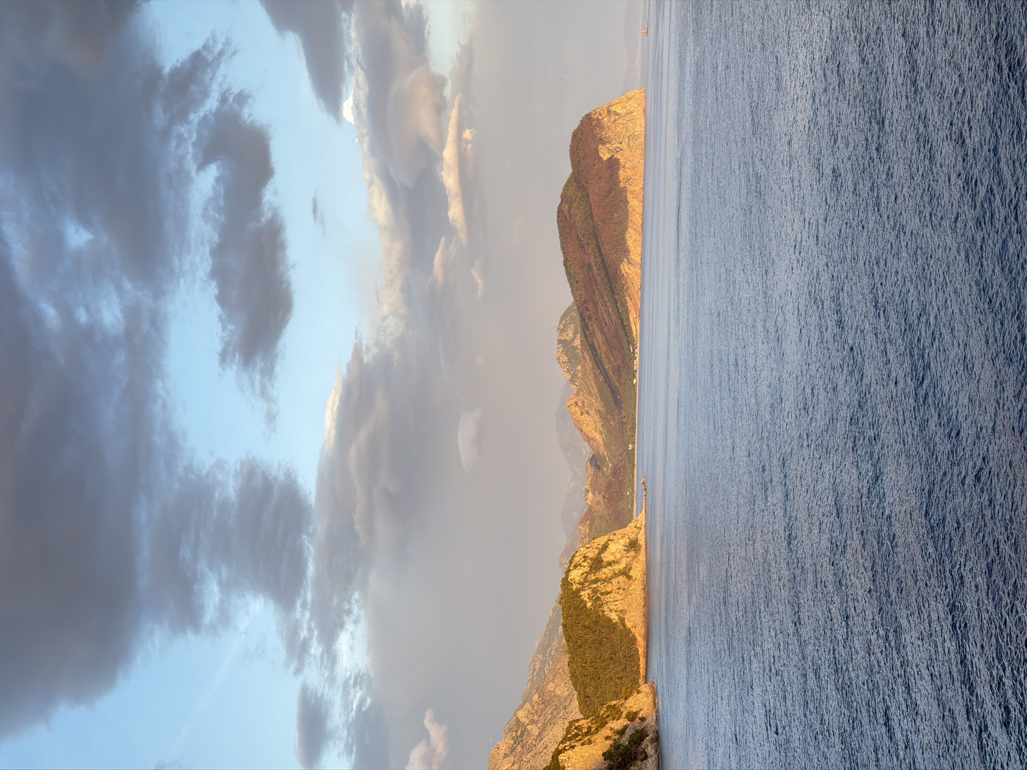 The image depicts a coastal landscape, likely at or near Mirište, Montenegro. The scene features a rocky coastline that appears to be bathed in the warm light of either sunrise or sunset. The rock formations exhibit steep cliffs and varied textures, colored in tones ranging from yellow to brown. The sea occupies a significant portion of the frame, showing a slightly choppy surface with an even, blue hue.
Above the land, a sky is dominated by dark, heavy clouds on the left side of the frame, contrasting with areas of lighter sky. These clouds suggest the possibility of an approaching storm.