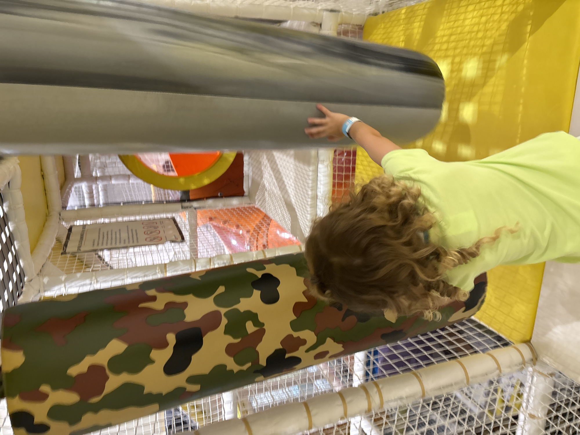A young child with curly blonde hair, wearing a neon green t-shirt and a blue wristband, is playing in an indoor soft play area. The child is viewed from behind, reaching out with their right hand towards a large, gray, cylindrical pillar. To the left of the pillar is a large, cylindrical object covered in a camouflage pattern of green, brown, and black. The play area features yellow padded flooring, white netted walls, and visible scaffolding above. In the background, glimpses of orange structures and a circular orange and yellow design can be seen, suggesting a jungle gym or activity zone. The scene is well-lit, indicative of an indoor recreational facility. The location is an indoor play area in Dubai, United Arab Emirates.