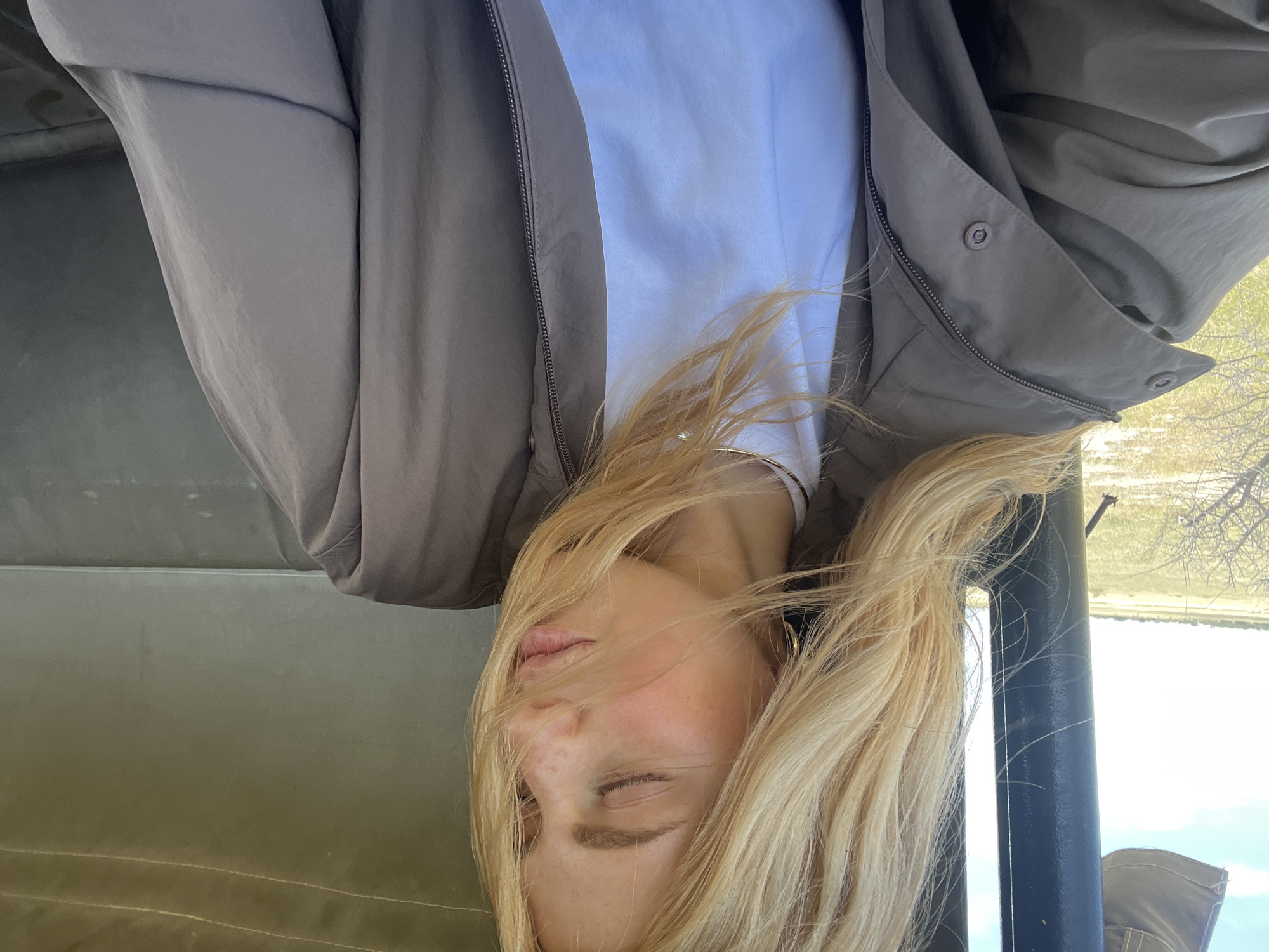 The image shows a woman with blonde hair, wearing a white t-shirt and a taupe-colored jacket, likely inside a vehicle, possibly a safari vehicle, with a canvas top. In the background, visible through an opening, is a landscape with trees and water, possibly indicating a safari or nature-based setting. Considering the mention of Gucta, Botswana, this is likely a safari experience within the vicinity of Gucta. The image is presented upside down.