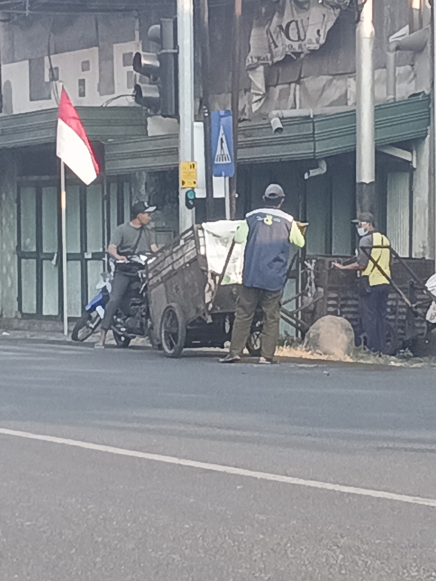 In Kemangsen, Indonesia, a person on a motorcycle with a trailer attached is parked on the side of a street. The motorcycle rider, wearing a dark shirt and a cap, is looking to the right. The trailer is laden with white material. Two other individuals are also present near the trailer. One, wearing a dark blue vest over a light shirt and khaki pants, is standing with their back to the camera. The other, wearing a yellow vest and a mask, is standing to the right of the trailer and appears to be interacting with it or the person in the blue vest. In the background, a building with a green awning and a faded banner overhead is visible. A traffic light and a pedestrian crossing sign are mounted on a pole. An Indonesian flag, red and white, is displayed on a pole in front of the building. The scene is set on an asphalt road with a white line marking the lane.