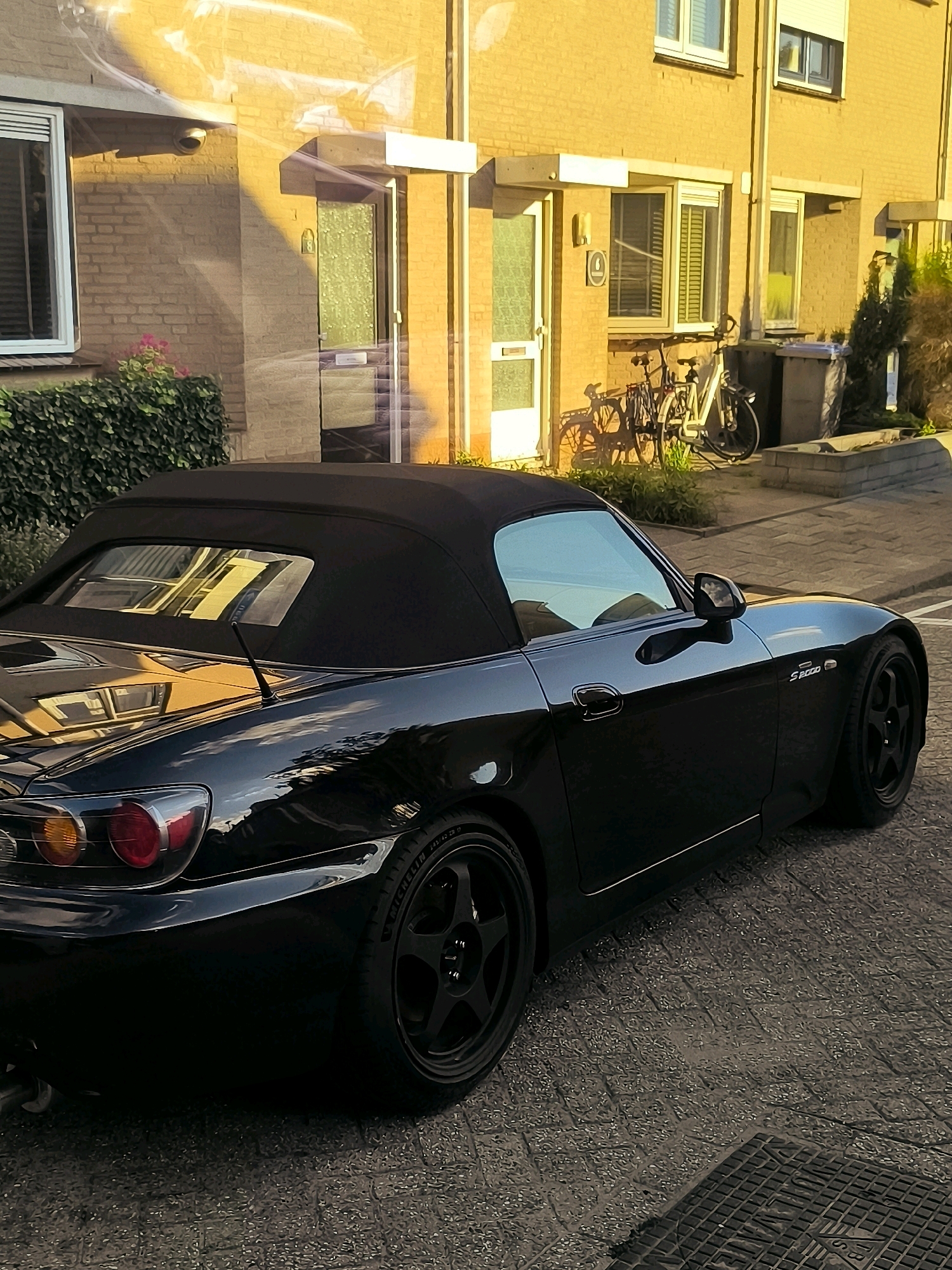 A black Honda S2000 convertible is parked on a paved street in front of a brick building in Oostvoorne, Netherlands. The car has its black soft top up. The building has yellow brickwork and features several windows with white frames and roll-down shutters. A green hedge runs along the front of the building. Two bicycles are leaning against the wall near a doorway. The street is paved with textured rectangular stones. The scene is illuminated by the warm, golden light of what appears to be sunset or sunrise, casting long shadows. The sky is visible reflecting on the car's polished surface.