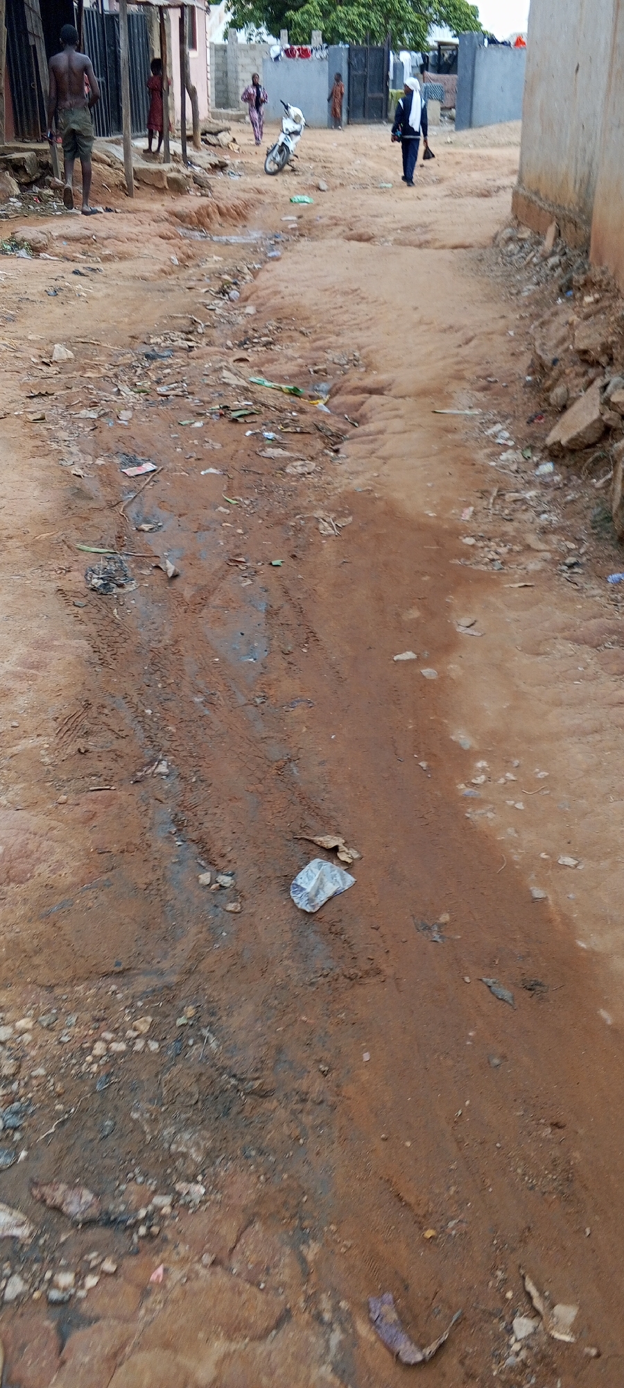 The image depicts a dirt road in Keffi, Nigeria. The road is uneven and muddy, with tire tracks visible. There is scattered litter, including plastic wrappers and debris. Several people are present in the background, including two children standing near a building, one person walking away from the camera on the left, one person walking in the road on the right, and others in the background. A white motorcycle is parked near the center of the image. Buildings of cinder block and sheet metal are visible, indicating a densely populated residential area. Laundry hangs on a line in the background.
