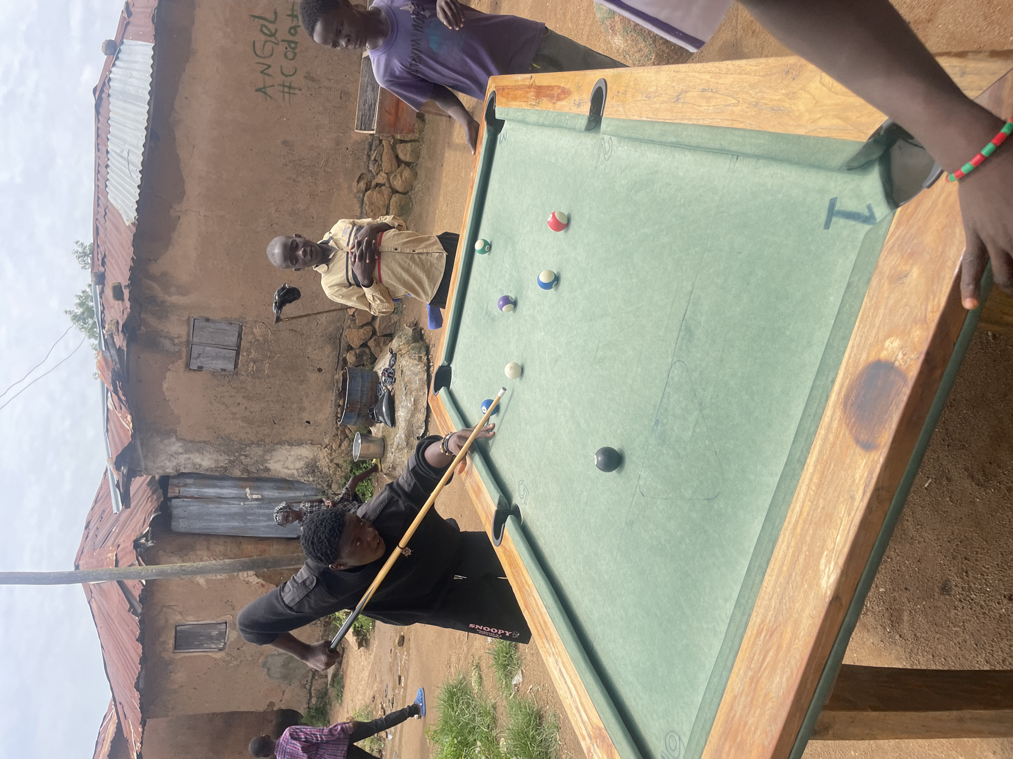In Jos, Nigeria, a group of young boys are gathered around a makeshift outdoor billiard table. The table has a green felt surface and a wooden frame, and is set up on the dirt ground. Several billiard balls are scattered across the table, including a black 8-ball, a striped blue and white ball, a solid purple ball, a solid white ball, and a striped red and white ball. One boy, wearing a black hoodie and sweatpants, is actively playing, aiming to strike a ball with a cue stick. Another boy in a tan shirt stands watching, his hands clasped in front of him. A third boy in a purple shirt leans against the table, observing the game. In the background, rustic, mud-brick houses with corrugated metal roofs line the dirt path. The sky is overcast, suggesting a cloudy day. The overall scene depicts a casual outdoor activity amidst a humble, rural setting.