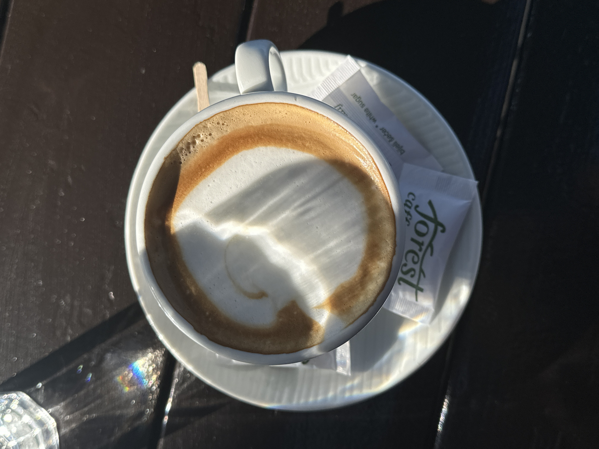 An overhead, close-up shot depicts a cup of cappuccino on a saucer at Forest Cafe in Šušanj, Montenegro. The coffee has a thick layer of foam with a light brown ring around the edge. A small wooden stirring stick is partially visible behind the cup, and two sugar packets rest on the saucer. Sunlight casts shadows and light patterns across the coffee and table. The table appears to be dark brown and made of wood. Rainbow colored glare can be seen next to the coffee cup.
