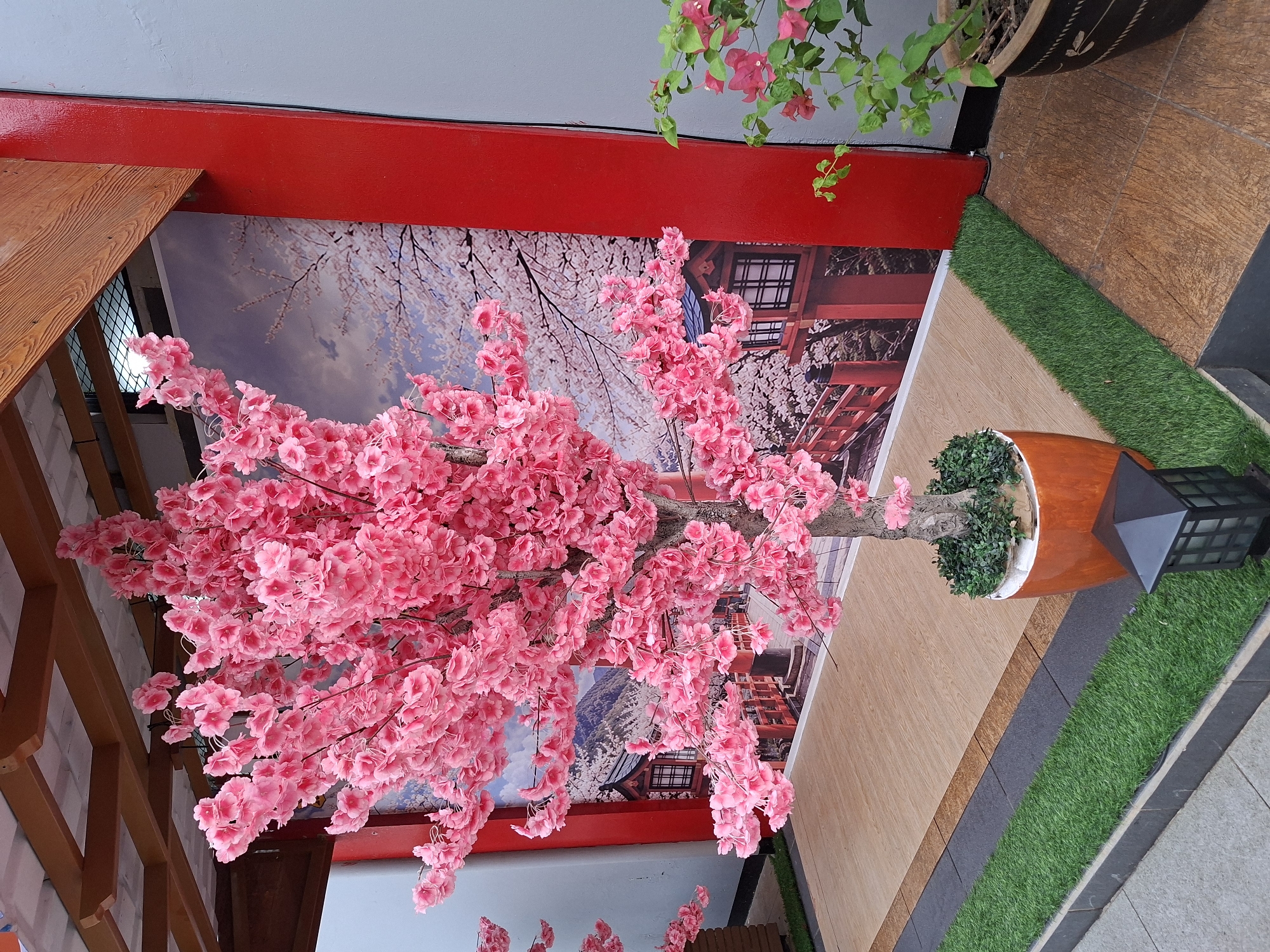 A life-sized artificial pink cherry blossom tree stands prominently in a paved outdoor area, possibly a garden or pathway in Surawangi, Indonesia. The tree is in full bloom, with a dense canopy of vibrant pink flowers. It is planted in a large, orange, tapered pot with dark green foliage at its base. Behind the tree, a mural depicts a traditional Japanese scene with a pagoda, a bridge, and more cherry blossom trees under a cloudy sky. The mural is framed by bright red architectural elements and a gray wall on the right. To the right of the mural, a small real plant with pink flowers in a dark pot is visible. The ground is a combination of light brown wood-look tiles and dark gray tiles with a strip of artificial green grass running along a pathway. A black, square lantern sits on the grass near the bottom right corner of the frame. The scene appears to be well-lit, suggesting it is daytime. There are no people visible in the image.