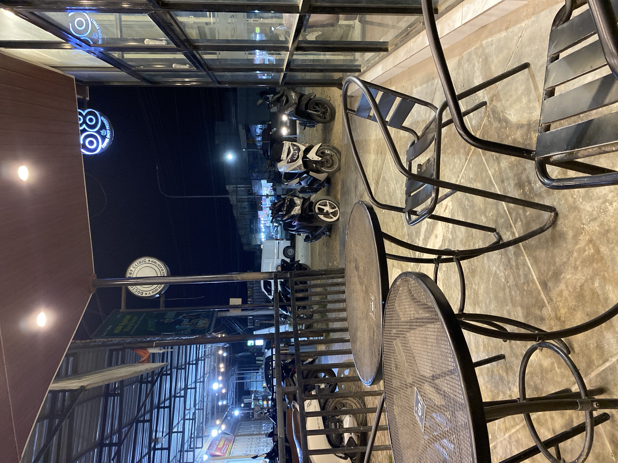 The photograph, taken at night, shows the exterior of a commercial establishment in Keudeublang, Indonesia. The scene is viewed from under an awning, with tables and chairs in the foreground. The furniture consists of metal-framed tables and chairs with a mesh design.
Outside the establishment, a row of motorcycles is parked along the curb. Across the street, lights from other businesses illuminate the area, including signs in Indonesian script. The reflections of these lights are visible in the glass windows of the building.
A neon sign for a "COFFEE SHOP PREMIUM" is visible on the building.