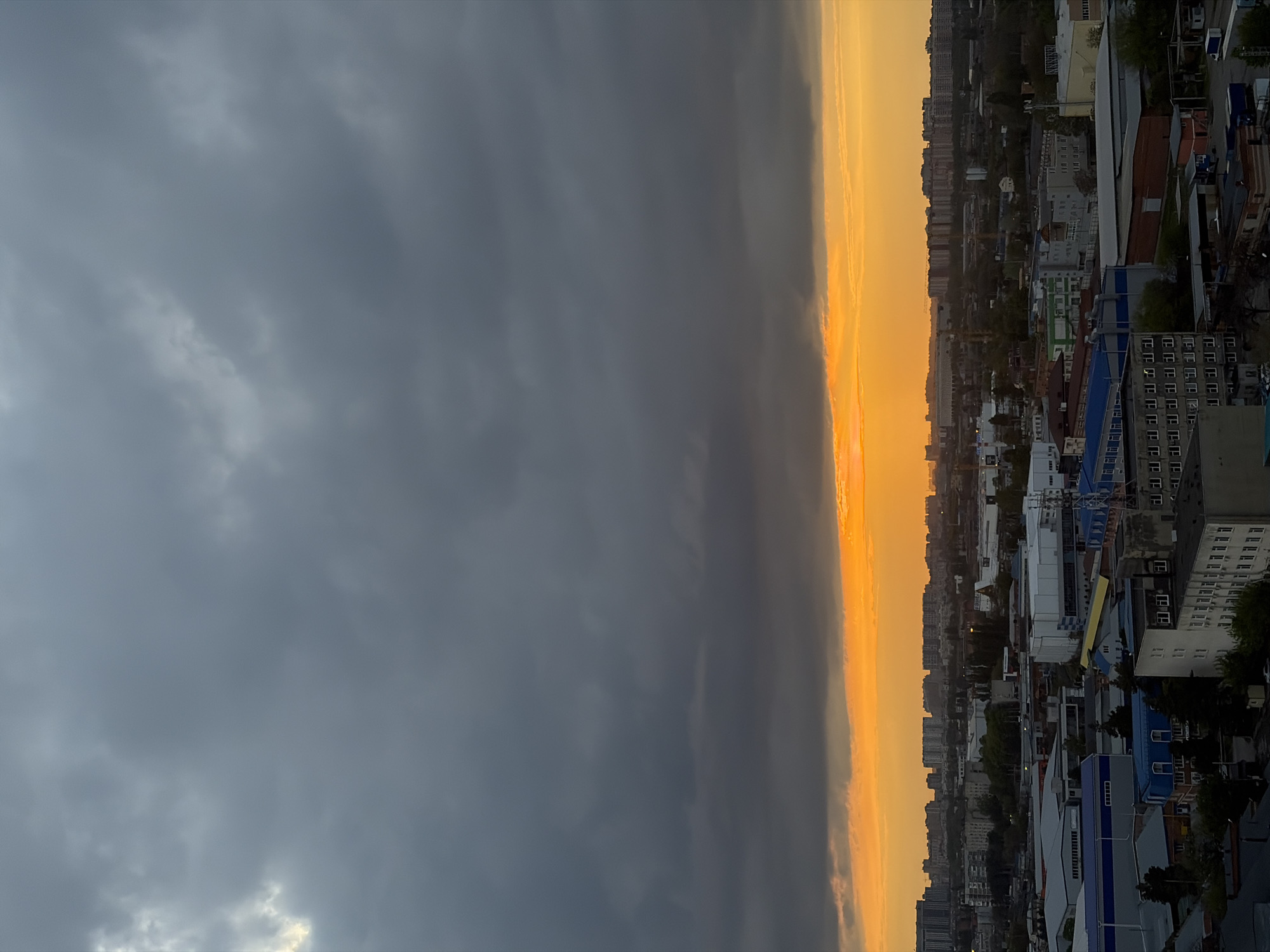The image depicts a cityscape in Krasnodar, Russia under a dramatic sky. Half of the frame is filled with dark, overcast clouds, suggesting an approaching storm. The other half shows a bright, golden sunset illuminating a dense urban area. Buildings of varying heights and architectural styles are visible, reflecting the warm light of the setting sun. Trees line some of the streets, providing a touch of greenery. The image is taken from a high vantage point, possibly from a tall building, offering a panoramic view of the city. The light and weather conditions create a strong contrast, emphasizing the division between the calm sunset and the impending storm. No people are visible in the image.