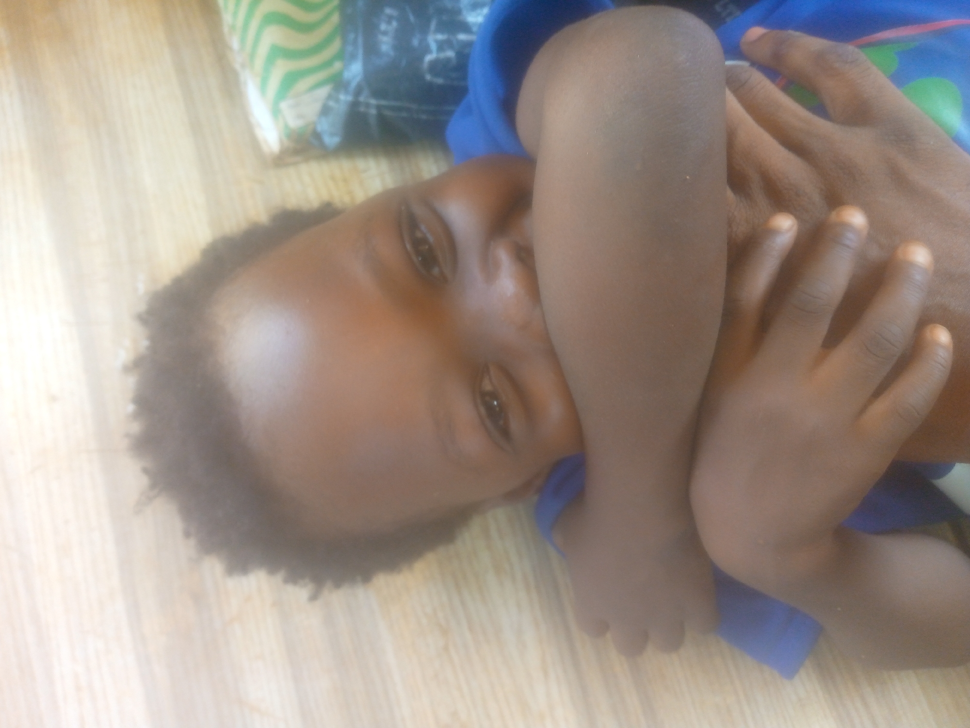 A close-up shot shows a young child lying on what appears to be a wooden floor, likely indoors. The child has dark skin and short, curly hair. The child's arms are crossed close to their face. The child is wearing a blue shirt. To the top left, there is a packaged object on the floor. The location is labeled as Unknown, Unknown.
