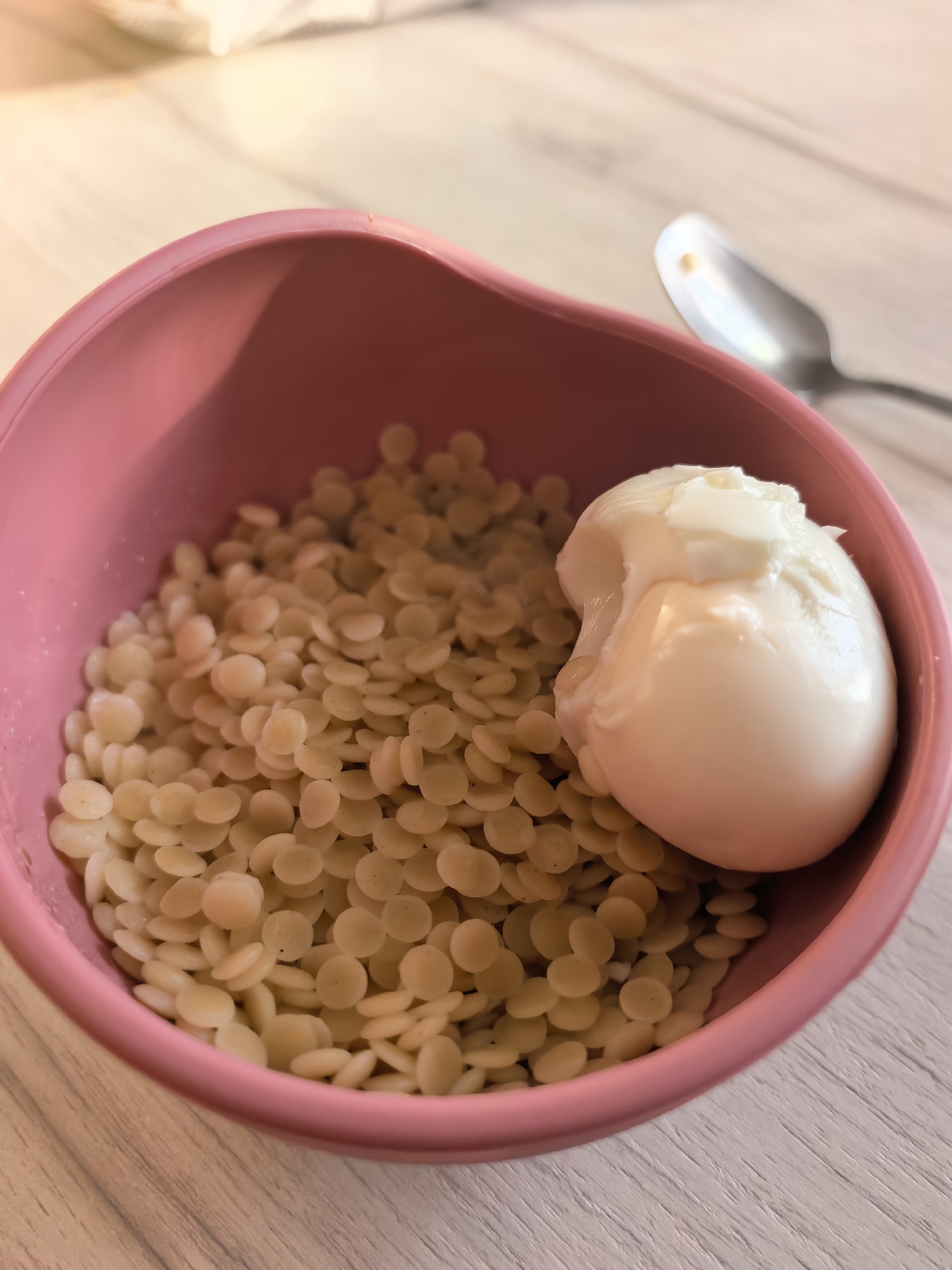 The image shows a pink bowl containing a pile of small, beige, disc-shaped objects and a peeled, soft-boiled egg with some of the egg white torn off. A silver spoon is partially visible in the background. The objects are sitting on a light-colored, wood-grained surface. The location of the image is unavailable.