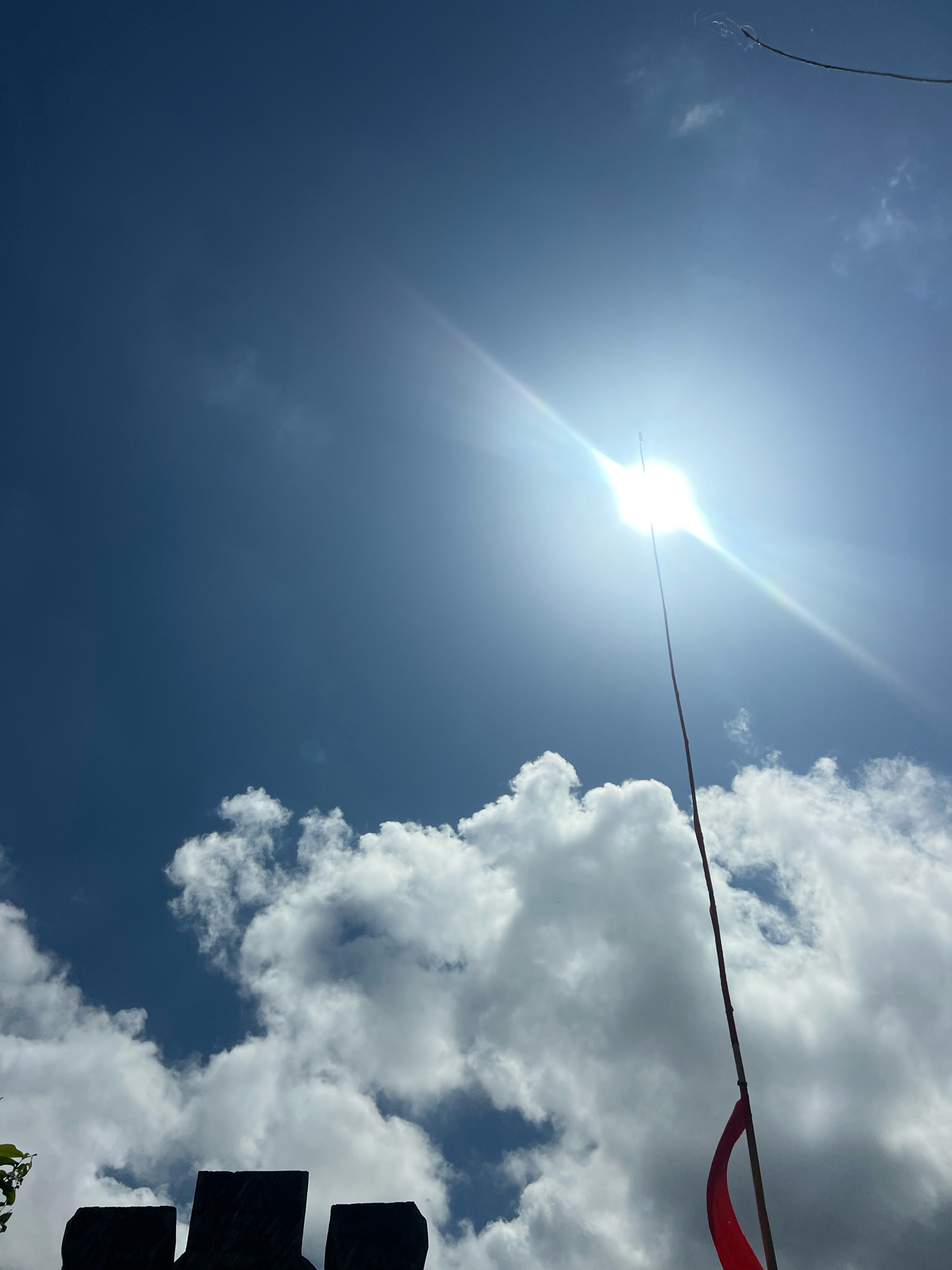 The image shows a bright, sunlit sky with fluffy white clouds against a deep blue background. The sun is shining intensely, creating a lens flare effect. In the foreground, silhouetted against the sky, are the crenellated tops of what appear to be stone battlements. A thin pole, topped with a small, bright red flag, extends upwards from the right side of the frame, positioned in front of the sun and clouds. The overall impression is of a clear, sunny day, possibly in a historical or scenic location.
This specific view, featuring stone battlements and a prominent flagpole with a red flag against the Denpasar sky, is characteristic of the **Bajra Sandhi Monument** in Denpasar, Bali, Indonesia. The monument complex includes historical displays and open grounds, and its architecture often includes elements resembling fortifications. The clear, bright sky is typical of Bali's tropical climate.
